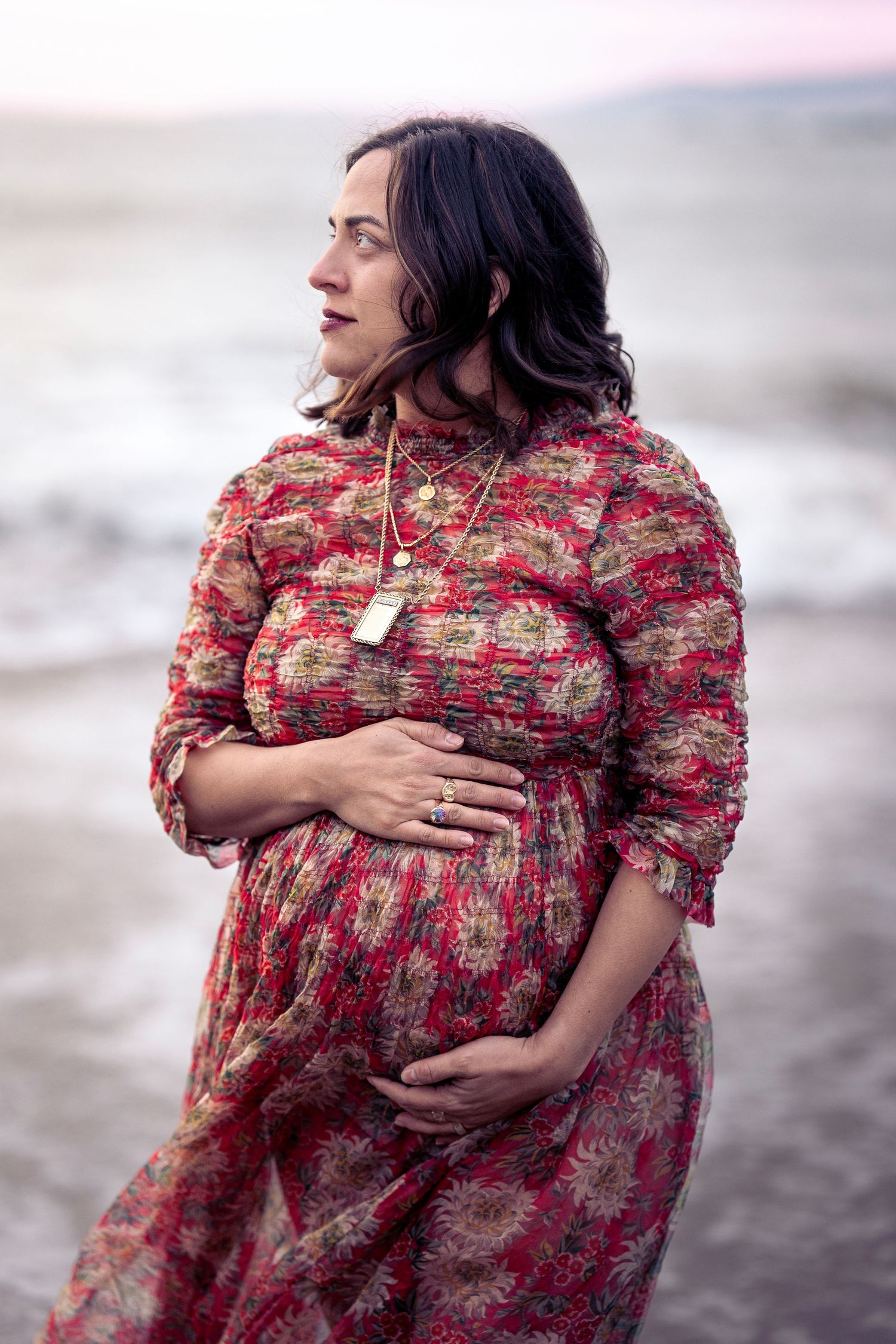 Someone in a flowing red dress stands at the beach during golden hour while touching their pregnant belly.