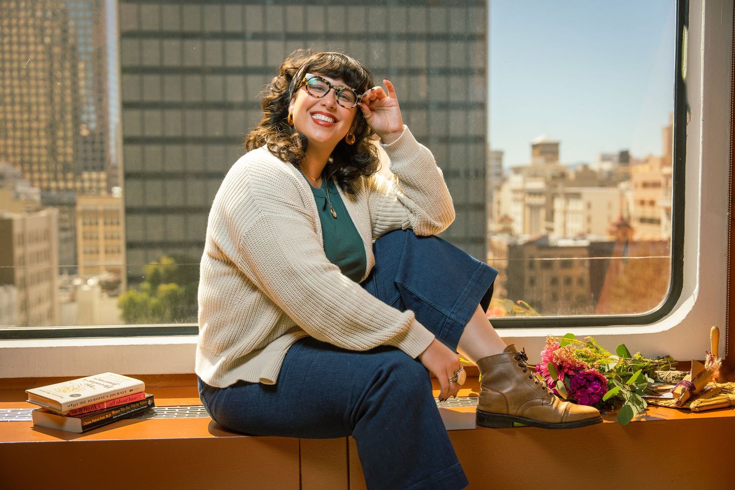 Someone in casual attire sits by a window with cityscape views, flowers and books nearby.