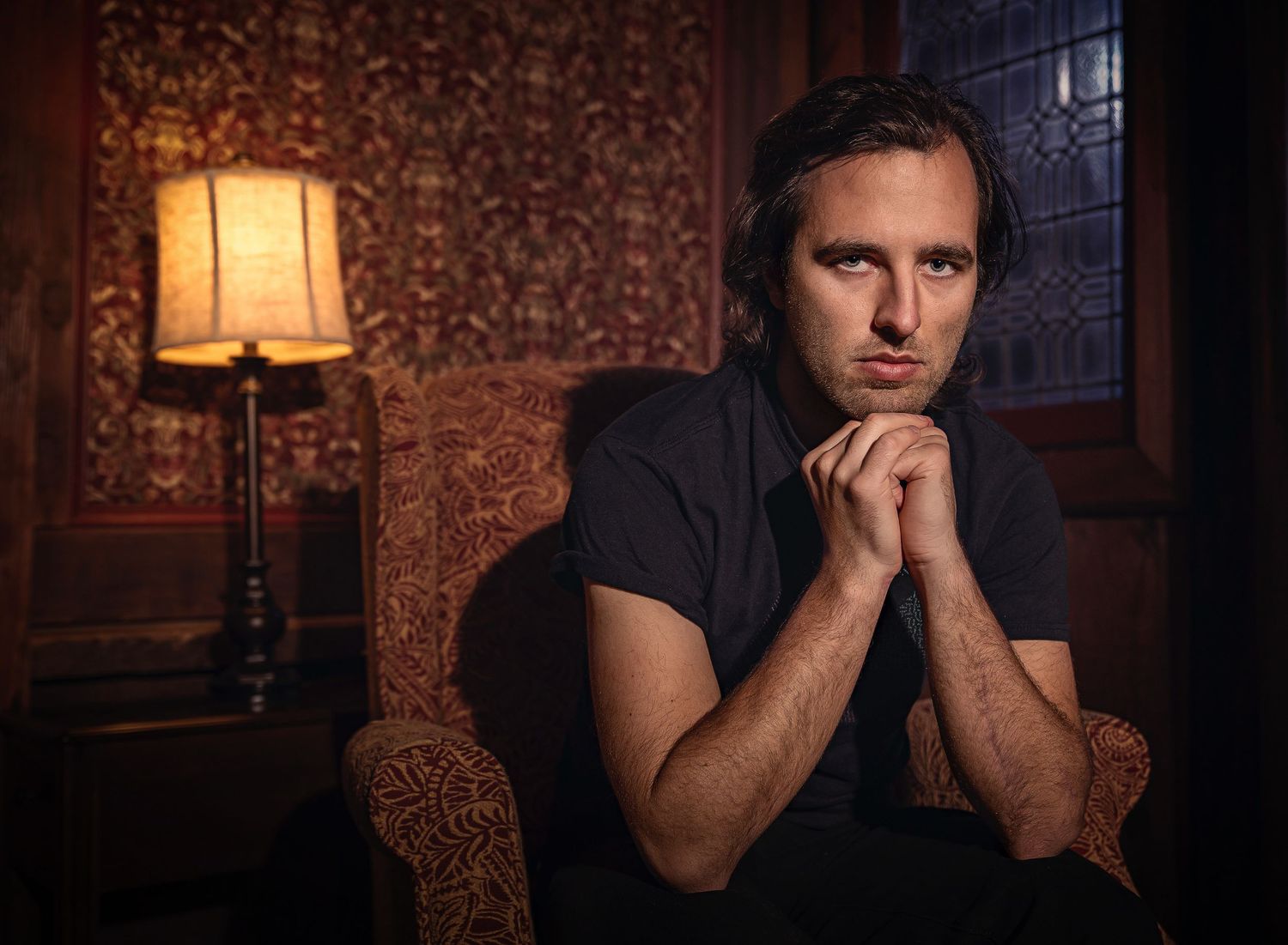 Moody portrait of someone sitting in a vintage chair next to a lamp in a dimly lit room.