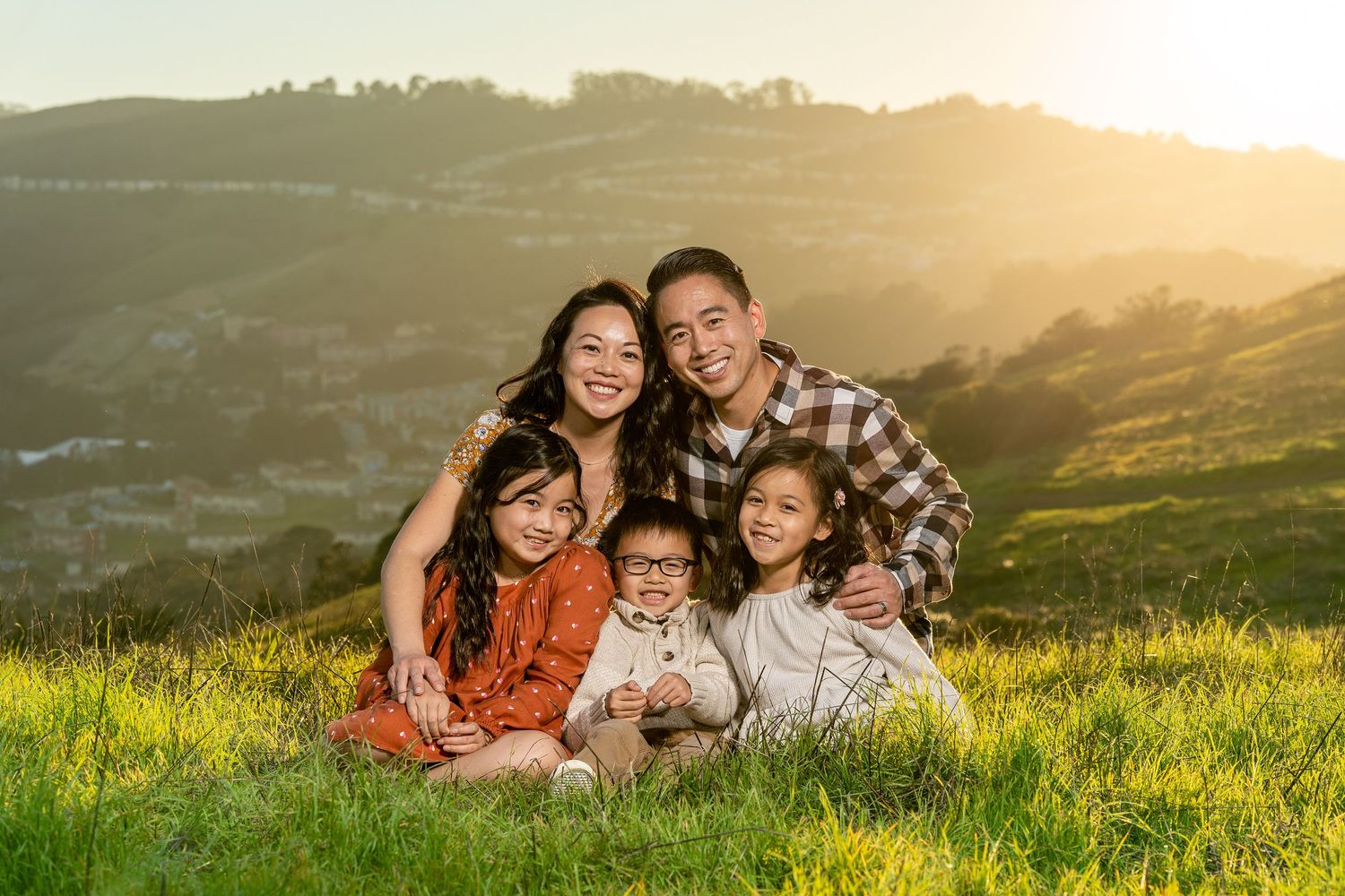 A joyful family sits together in golden sunlit grass on a hillside overlooking a scenic valley at sunset.