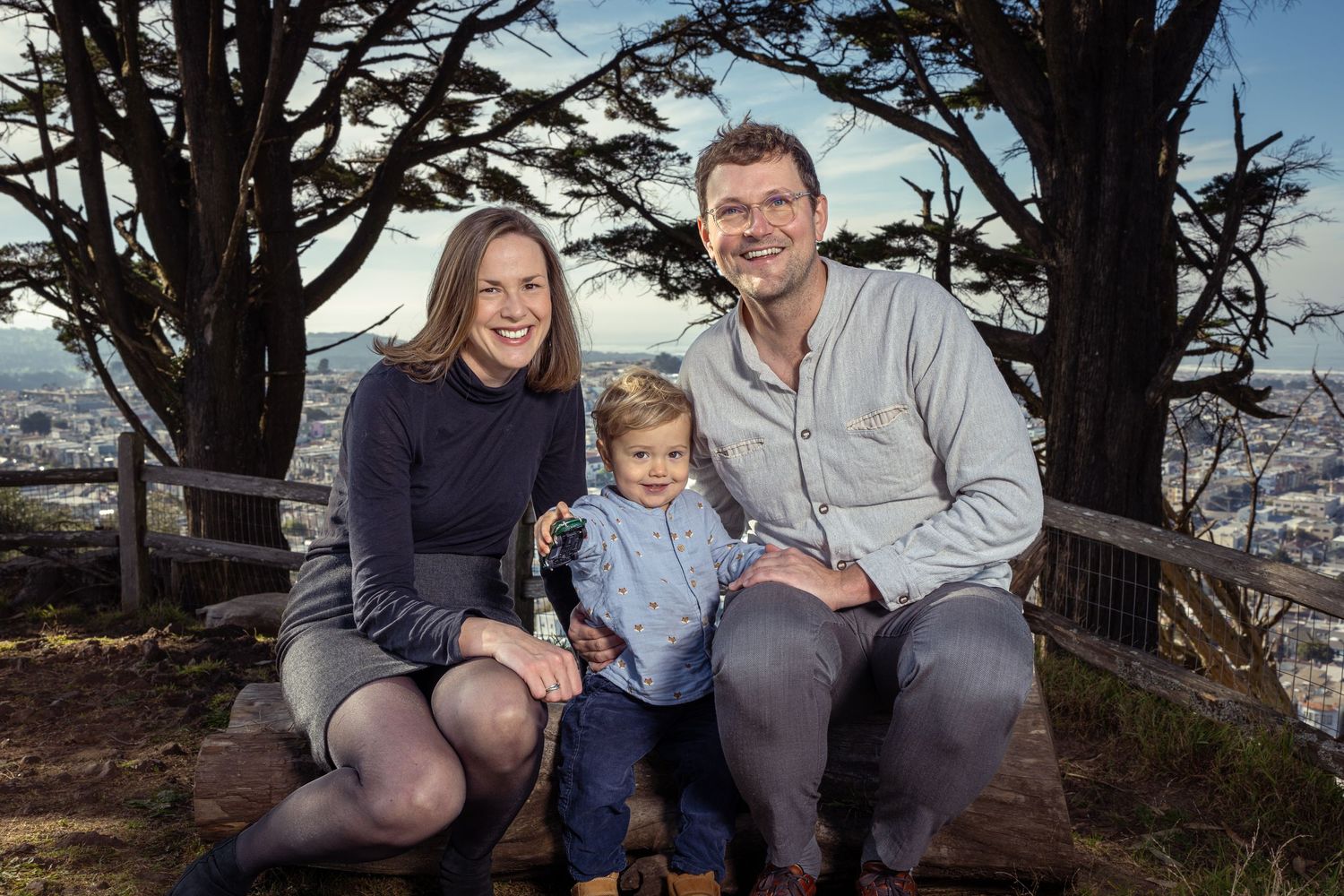 Family sitting together on wooden bench with city vista in background.
