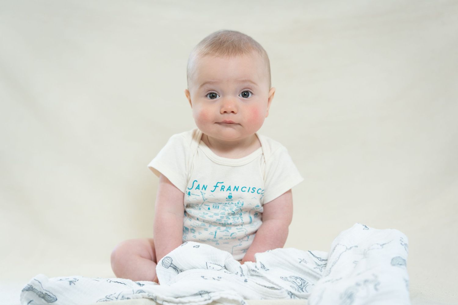 Baby wearing light blue graphic t-shirt sitting on white blanket.