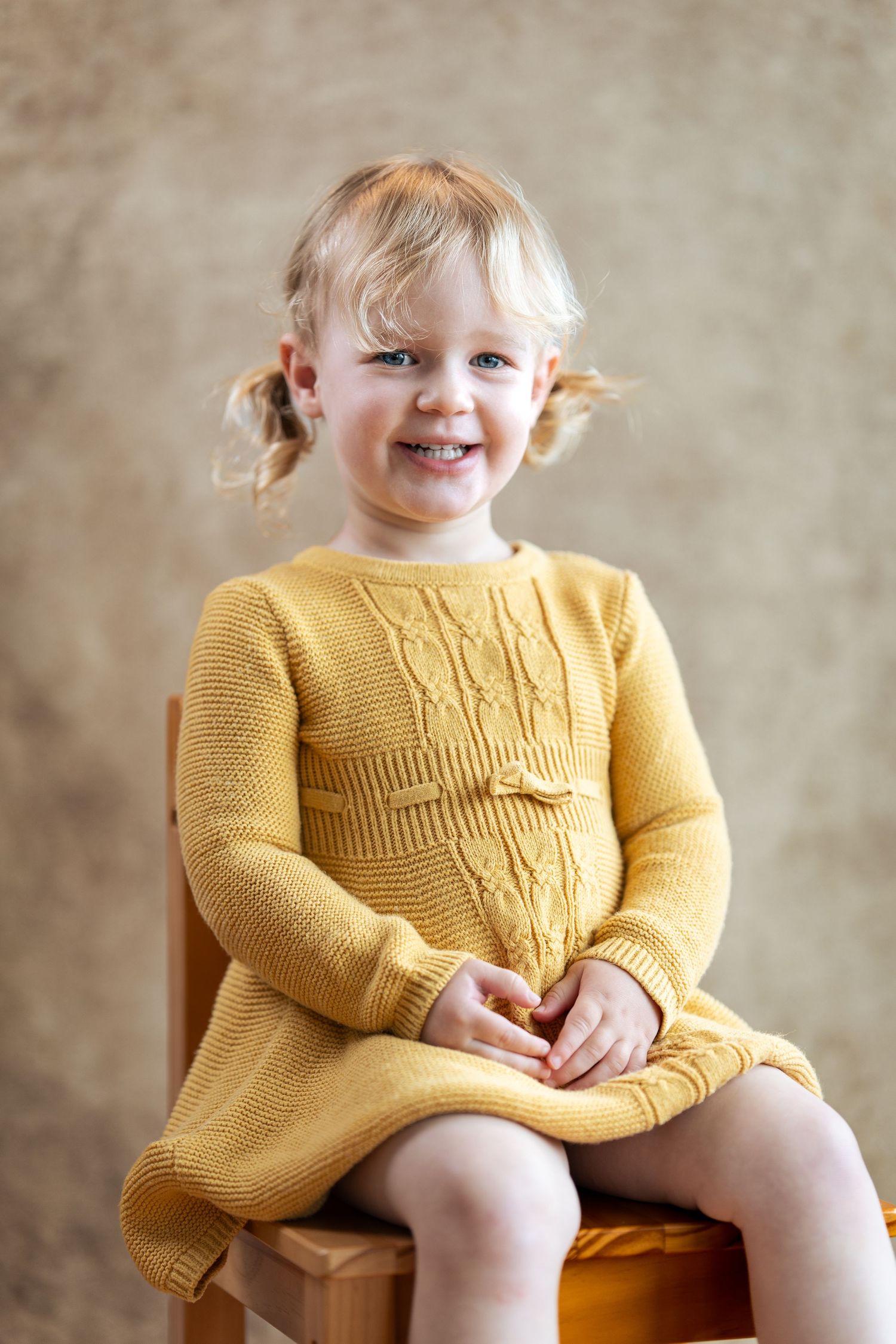 A child in a yellow cable knit dress sits playfully on a wooden stool.