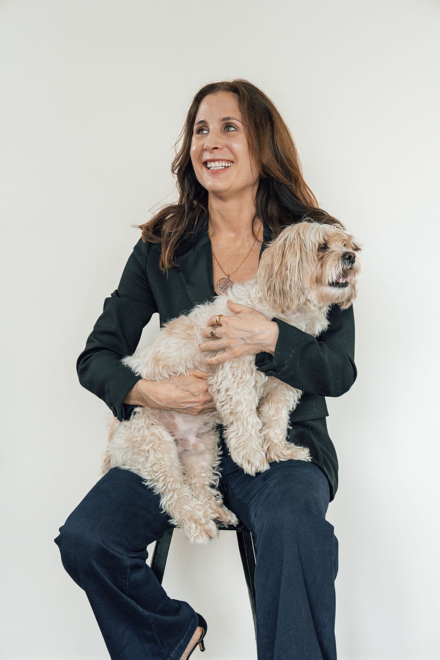 Someone in business attire holds a small cream-colored dog while posing against a white background.