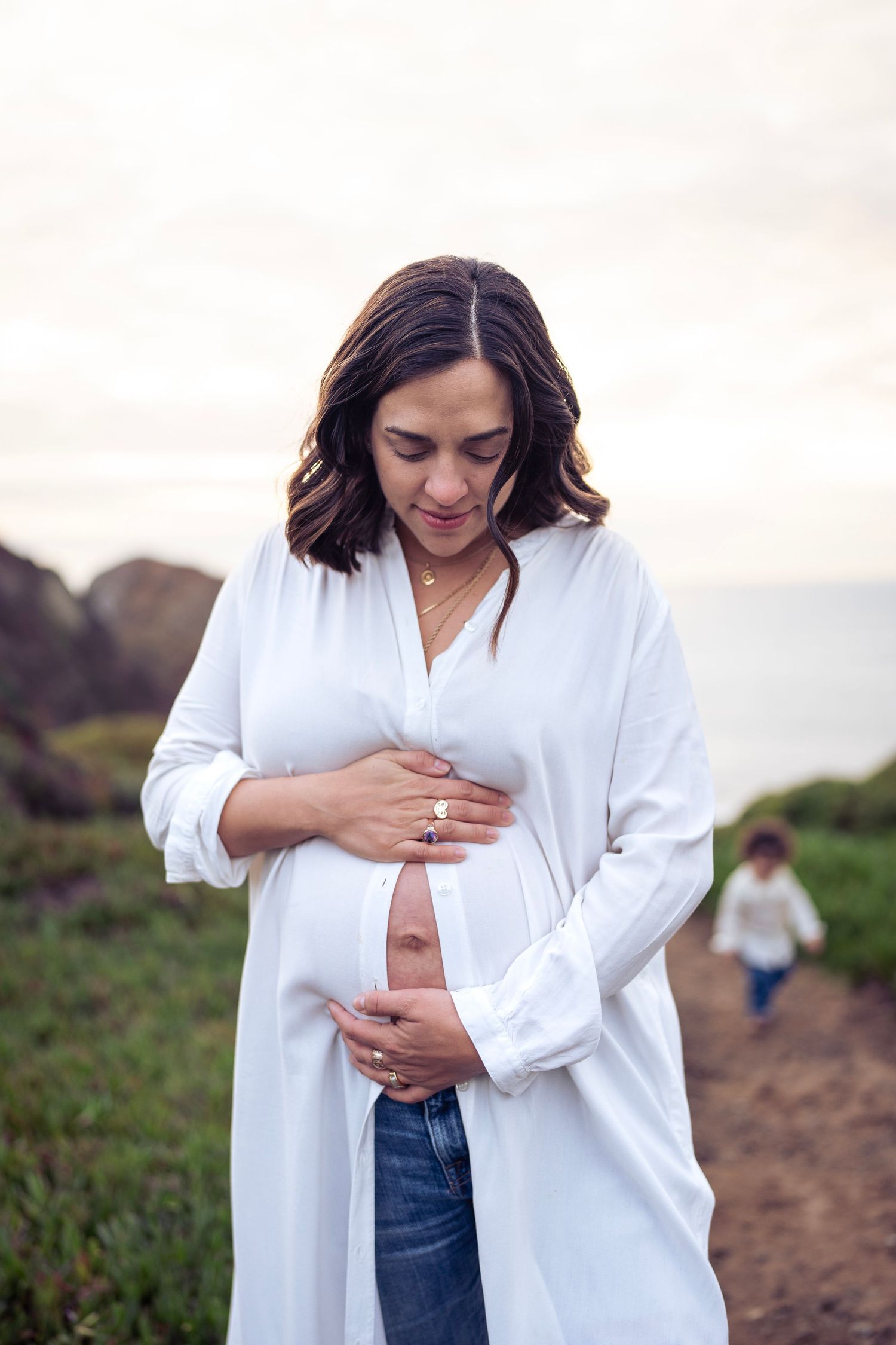 A casual maternity photo shows someone in a white button-down and jeans on a coastal path.