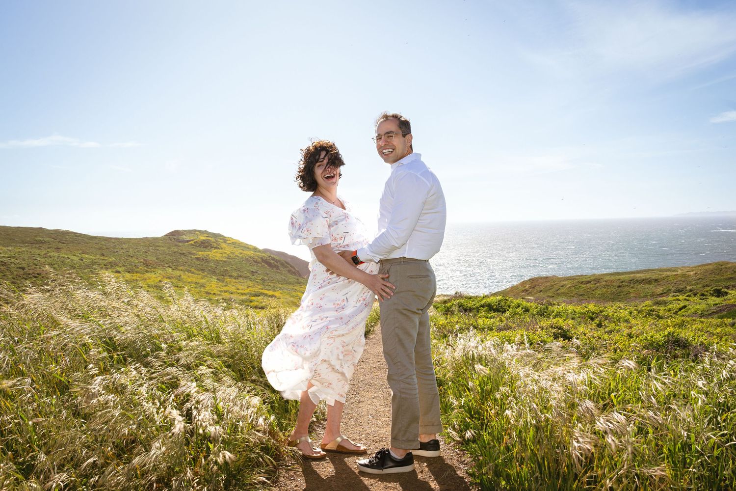 Couple in light clothing embracing on coastal path with ocean view.