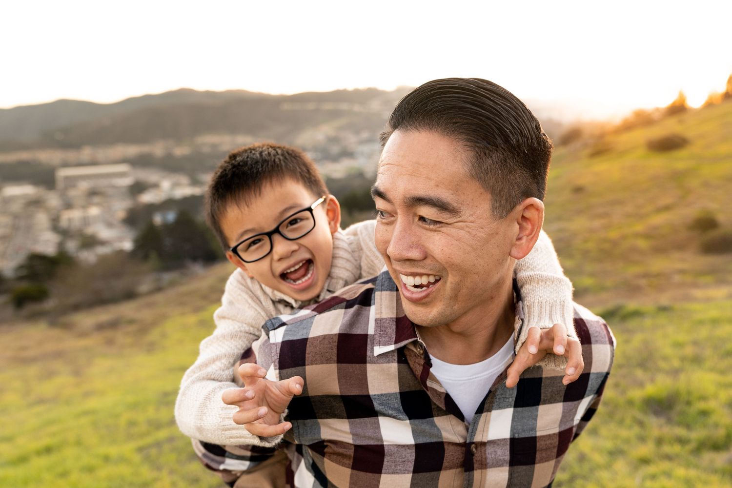 Father and child sharing joyful laughter during sunset on grassy hillside overlook.