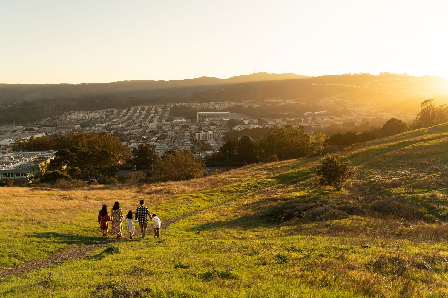 Silhouettes of people walking down a grassy hill at sunset overlooking a suburban landscape.