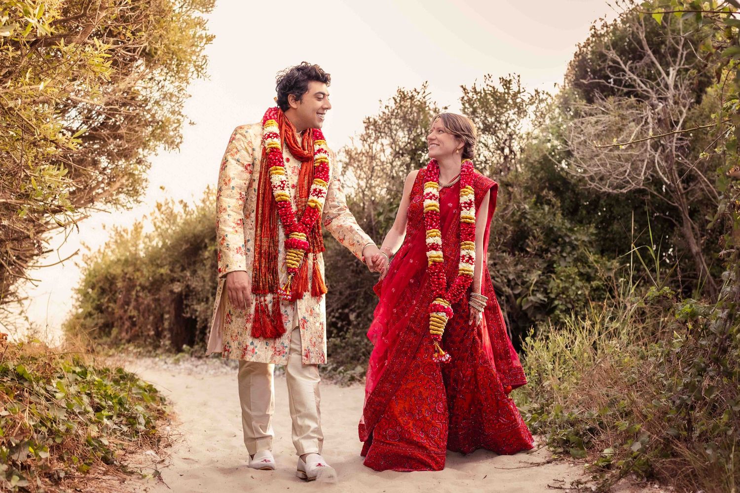 A couple in traditional Indian wedding attire walks along a path during golden hour.