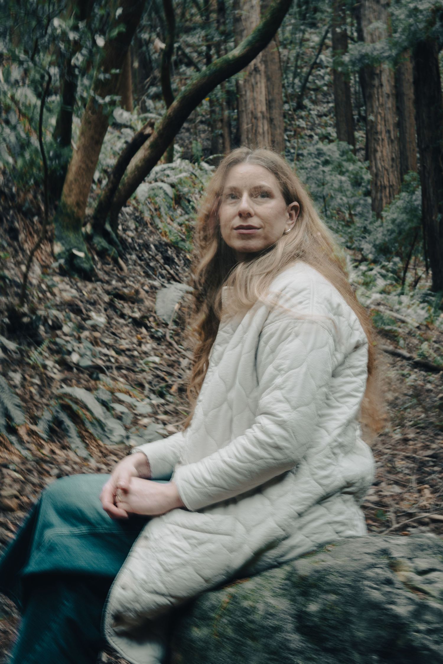 Someone in a white quilted jacket sits on a rock in a misty forest setting.