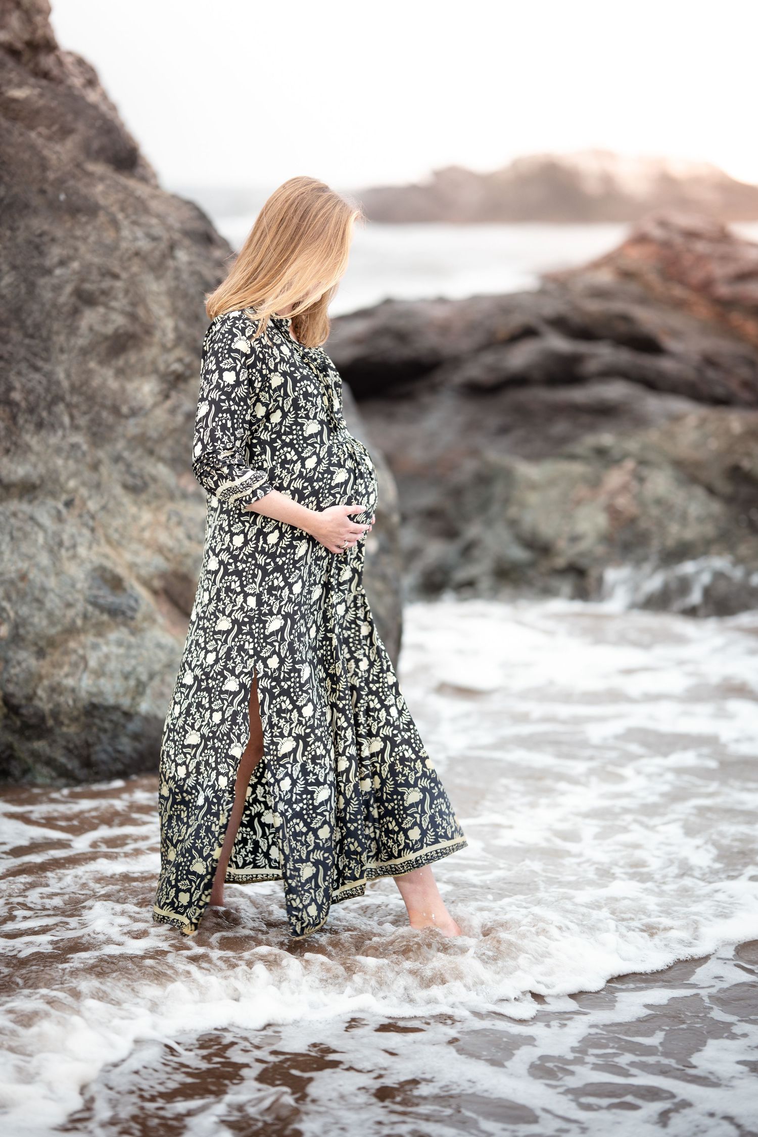 A person in a floral dress stands among ocean waves and rocks during a beach maternity photoshoot.