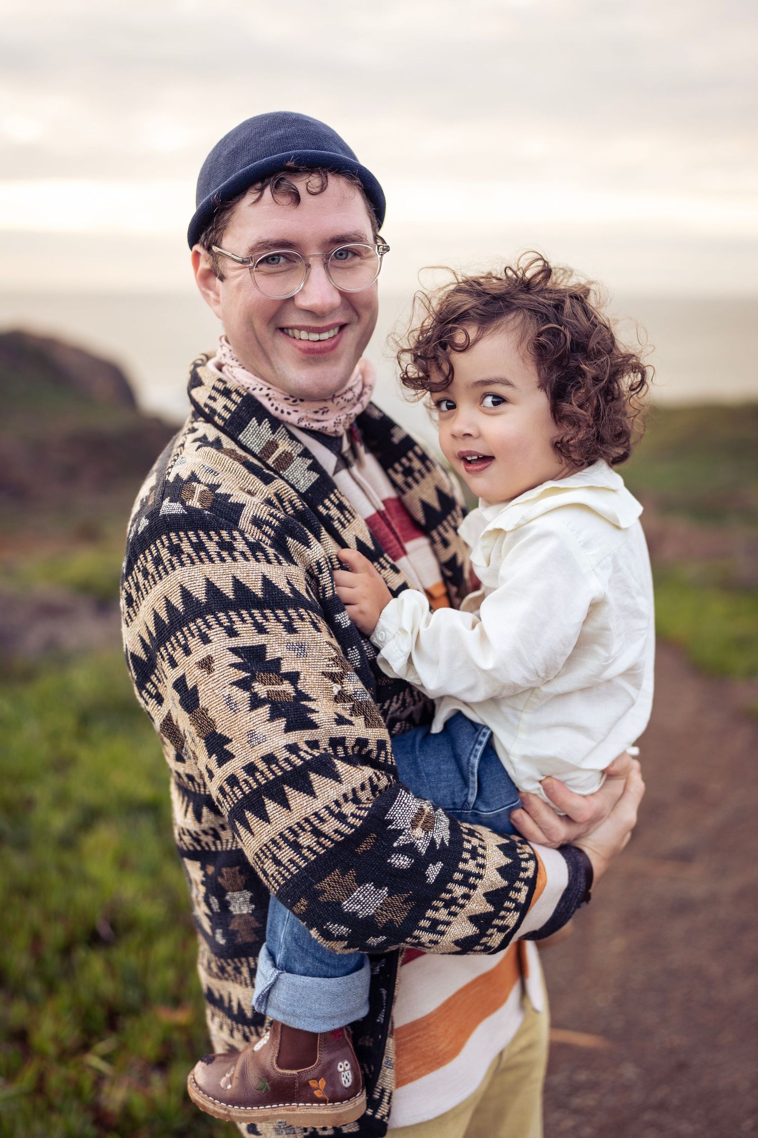 Parent in patterned sweater holding toddler outdoors by coast.