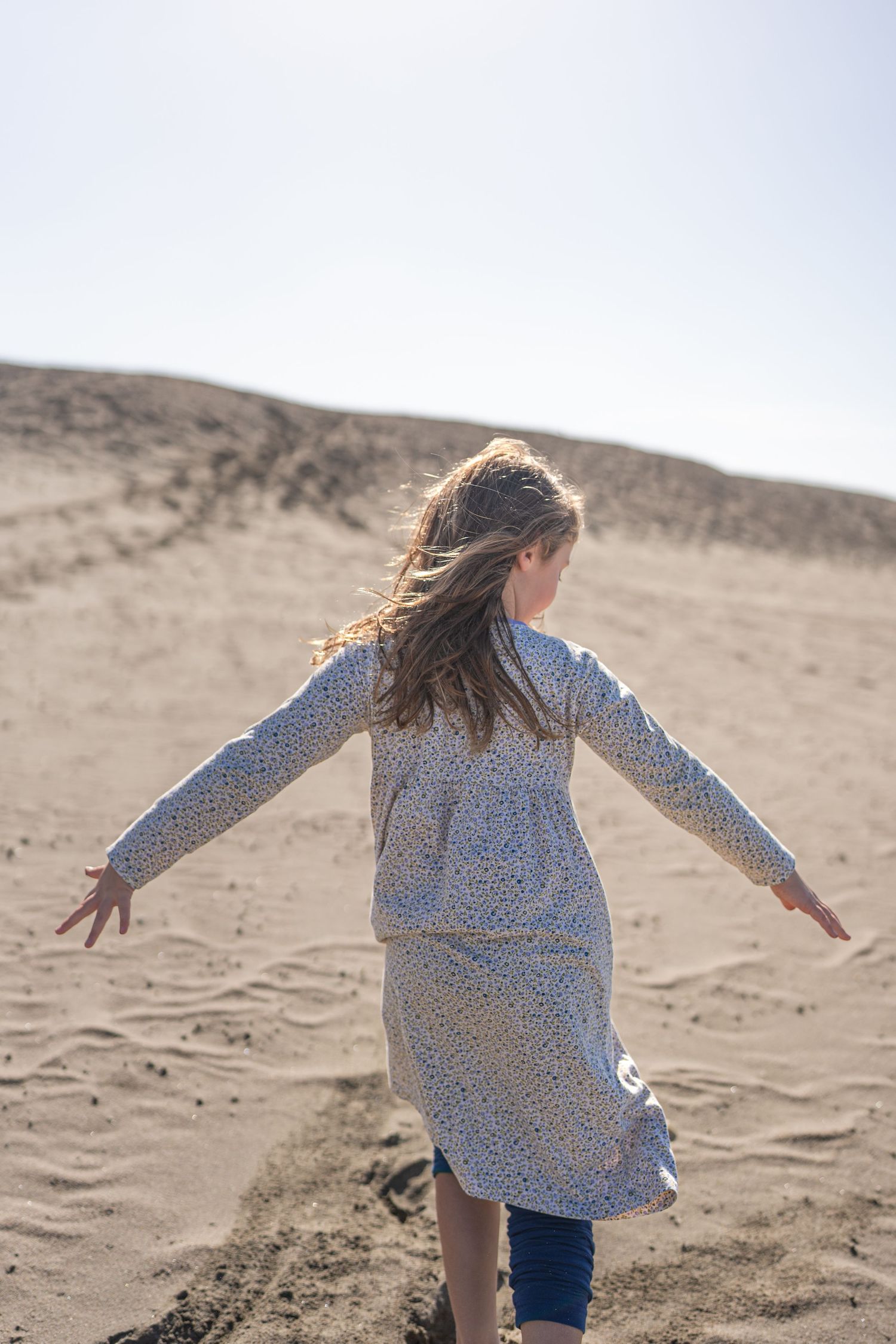 A person in a floral dress spins with arms outstretched on sandy dunes under a bright sky.