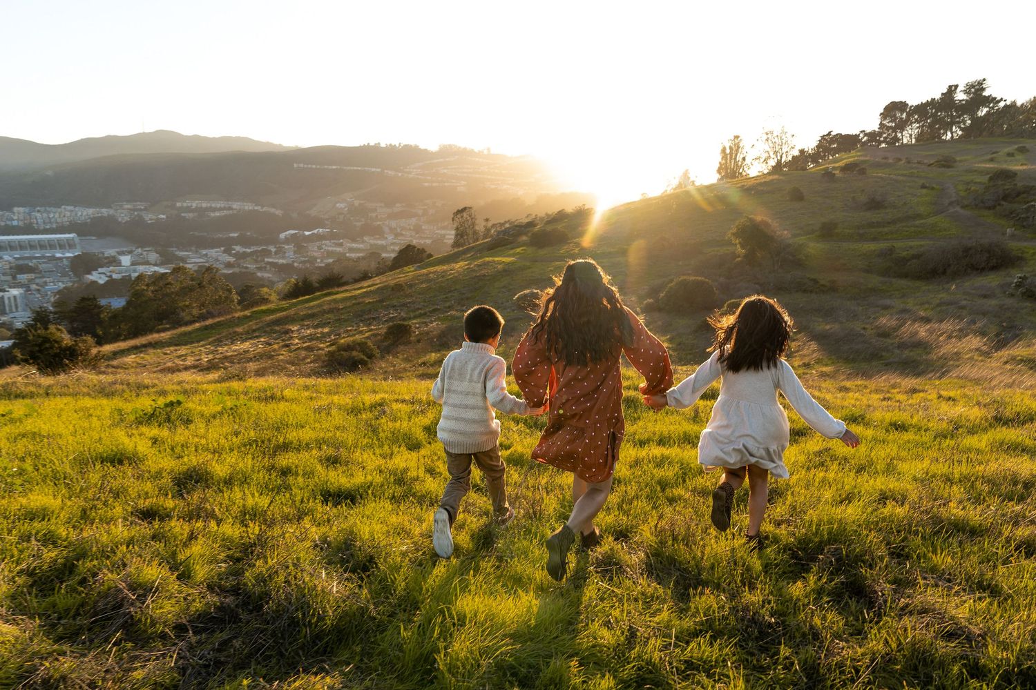 Three people run together through a sun-drenched grassy hillside at golden hour.