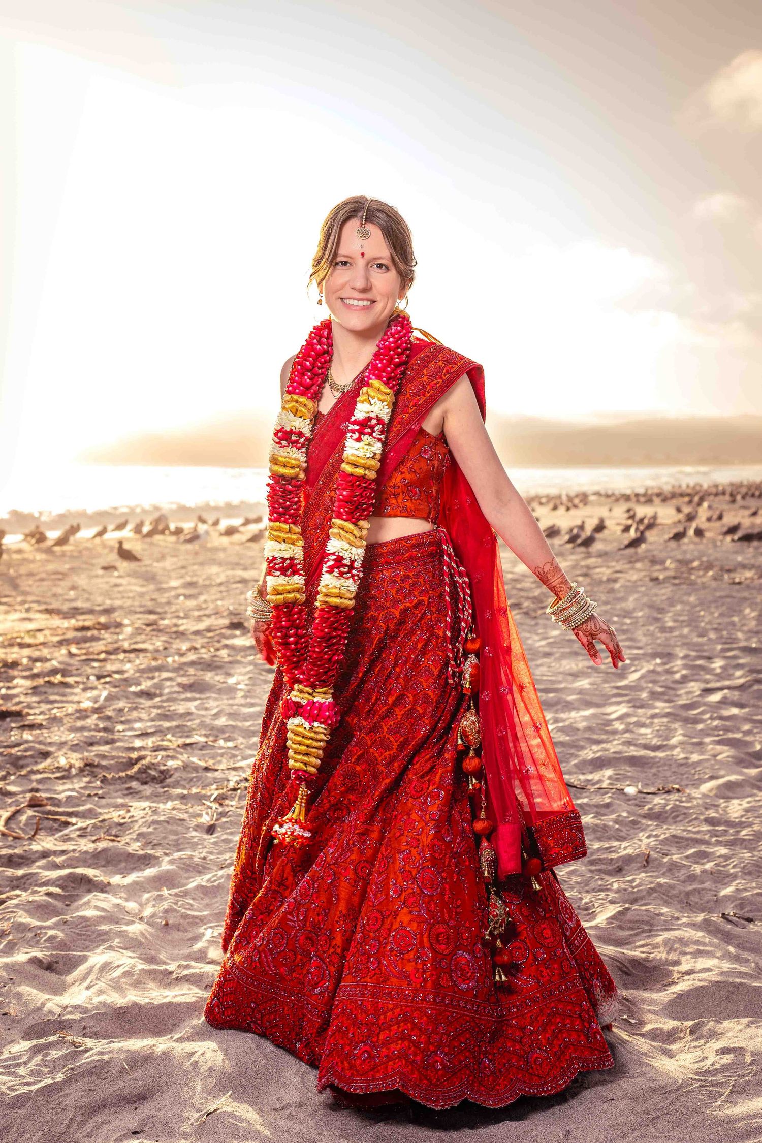 A woman in a traditional red Indian bridal outfit stands on a beach at sunset.