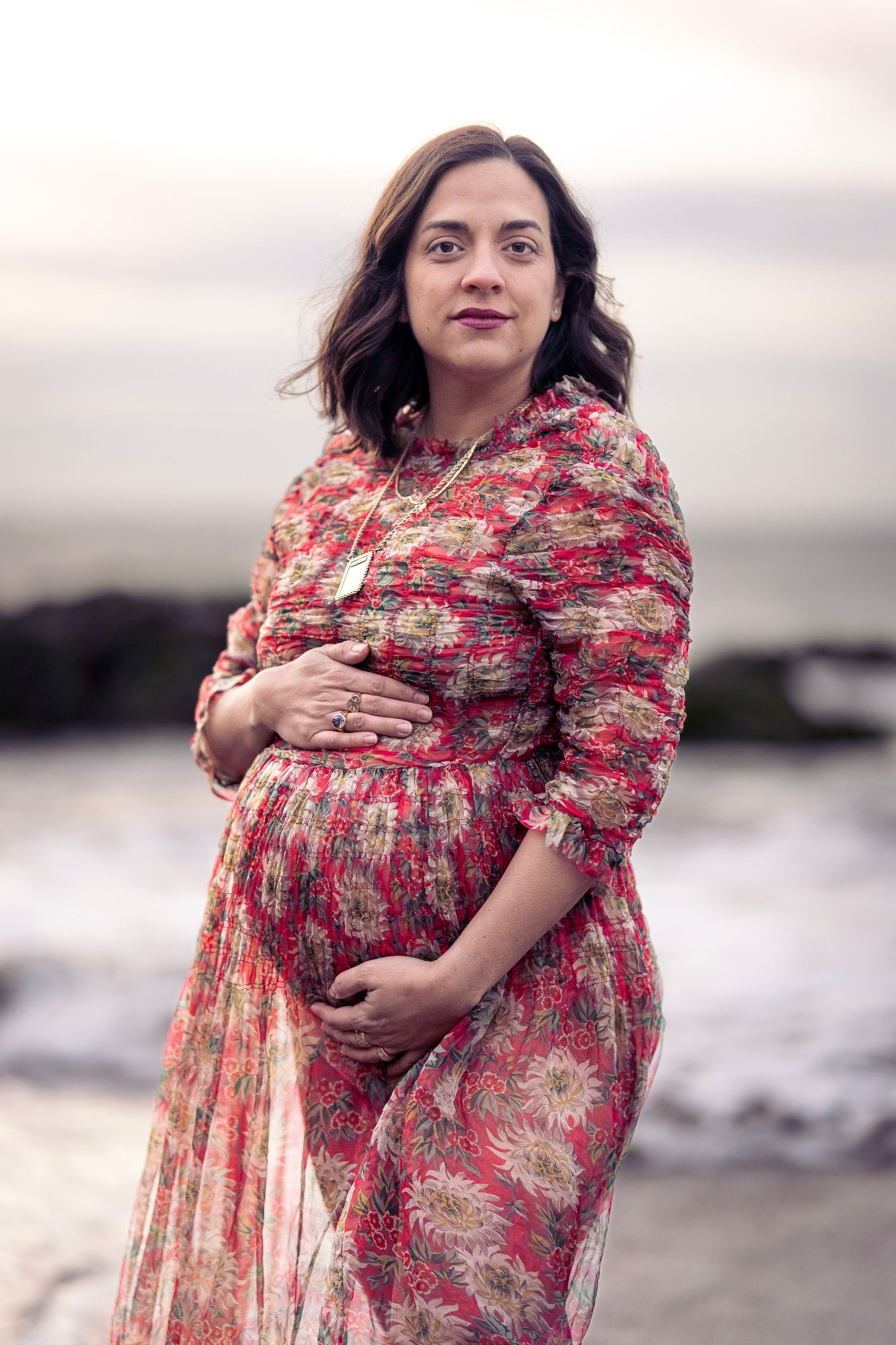 Expectant mother in floral dress poses on rocky beach during sunset.
