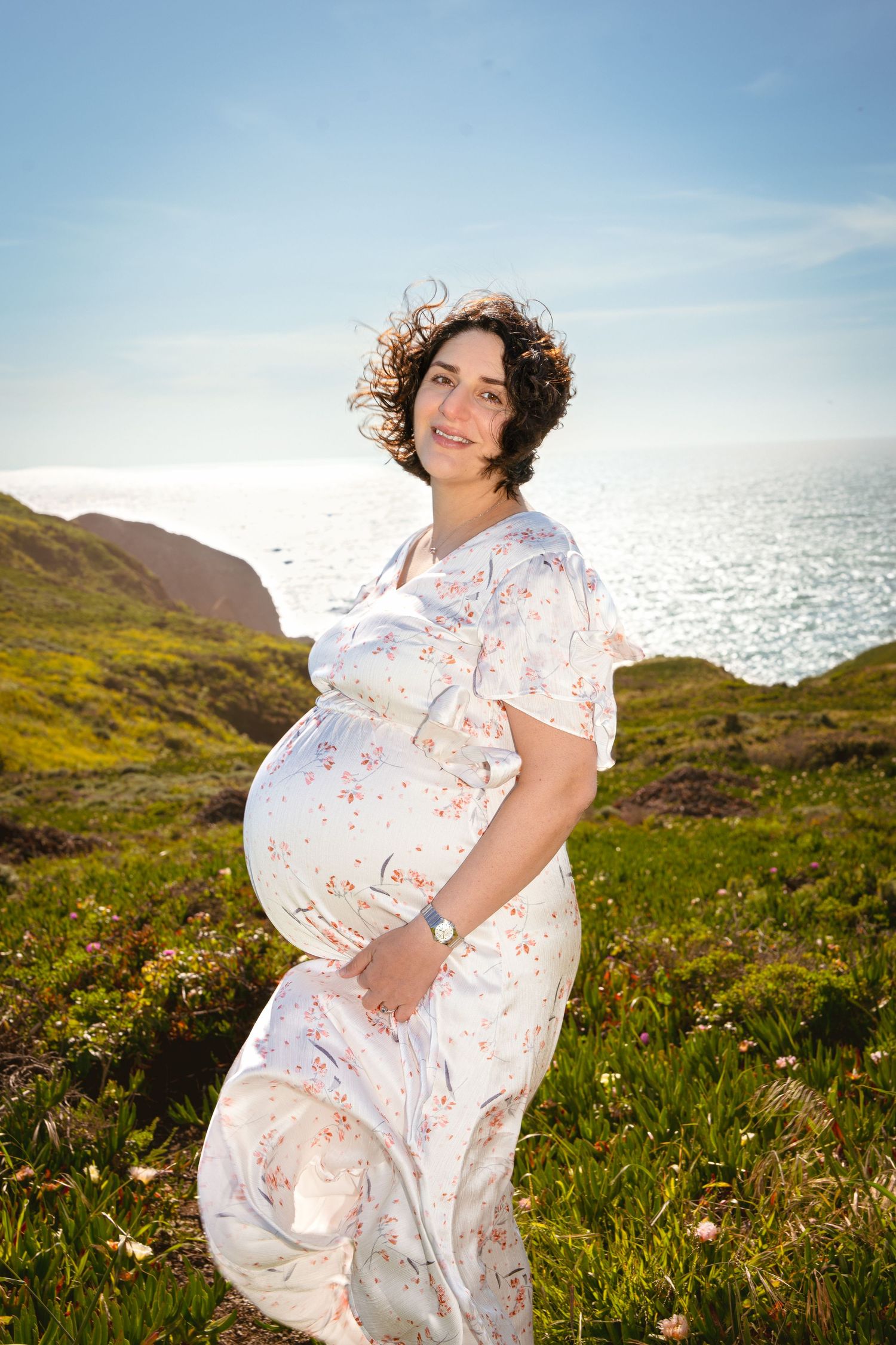 Expectant mother in white dress poses by coastline on sunny day.