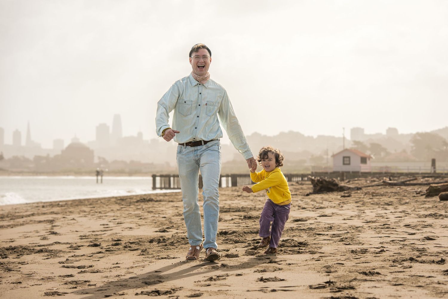 Two people joyfully running together on a sandy beach with a cityscape in the background on a misty day.