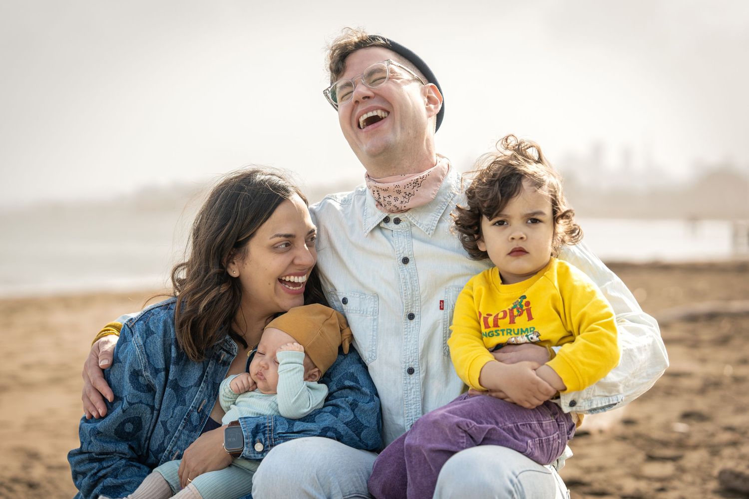 Happy family sits together laughing and smiling on a beach during sunset.