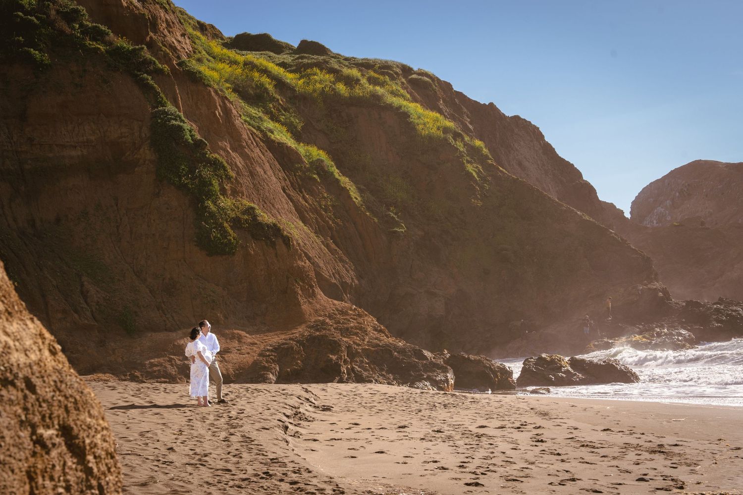 A lone figure walks on a sandy beach surrounded by towering cliffs and crashing waves at sunset.