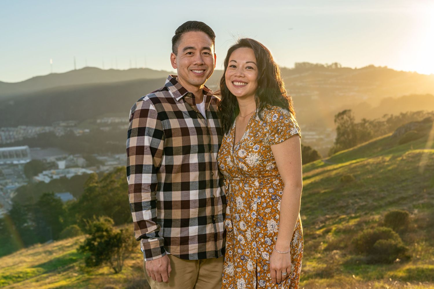 Couple in coordinated outfits embrace during golden hour on scenic hillside.