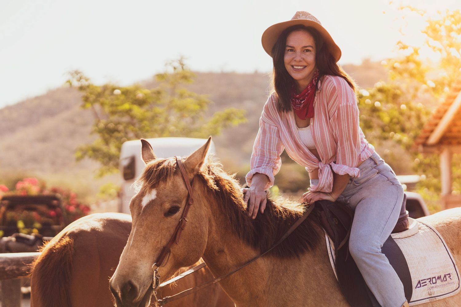 Person wearing a hat and pink shirt riding horseback in warm sunset lighting.