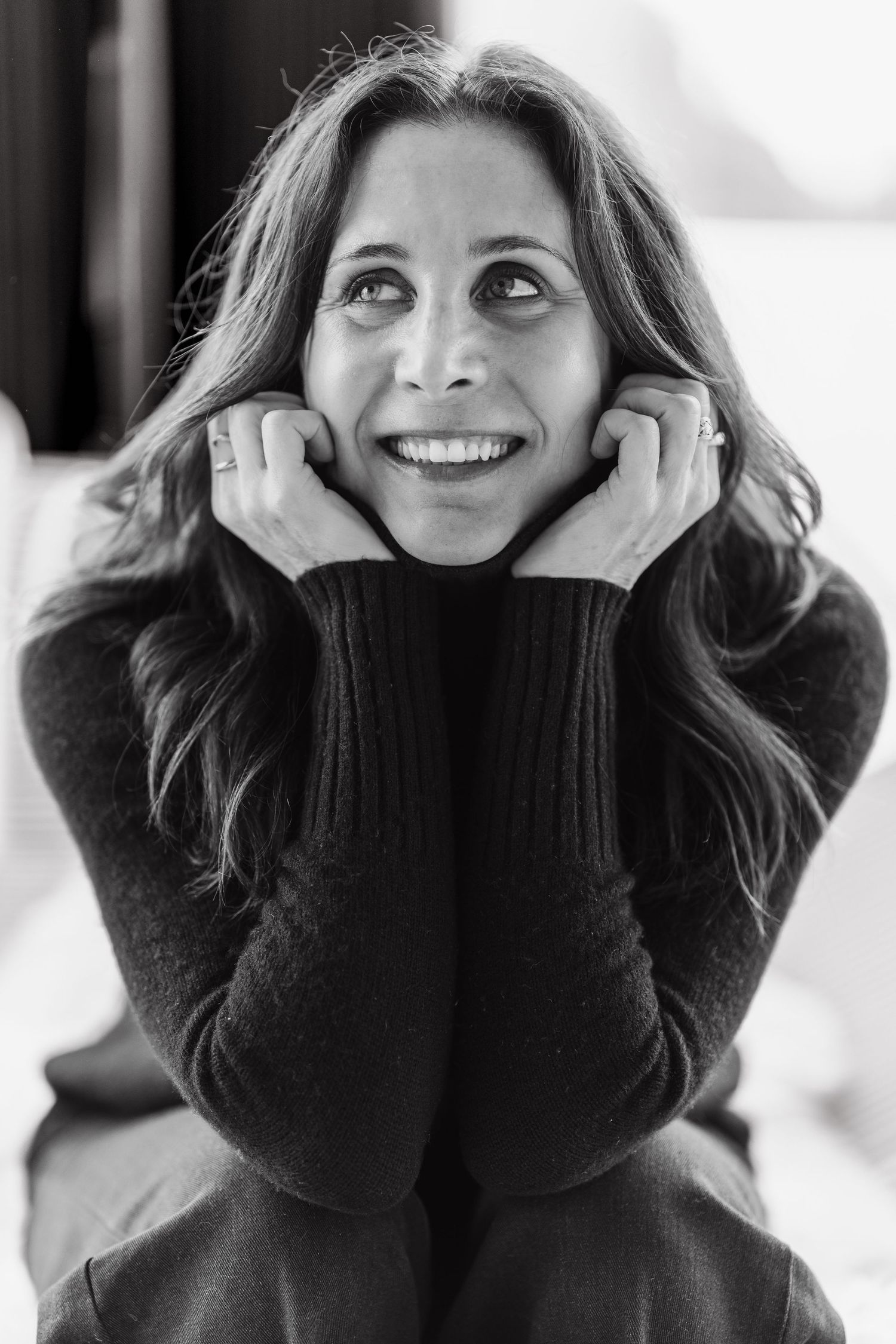 Black and white portrait of someone in a dark sweater smiling while resting their face in their hands.