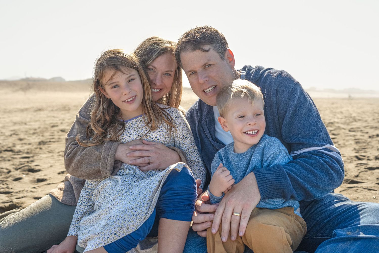 Family wearing coordinated blue and white outfits sharing tender moments together on a sunny beach.