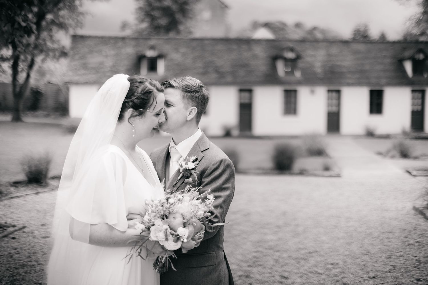 Black and white wedding photo of bride and groom sharing a tender moment, bride holding bouquet, rustic building backdrop.