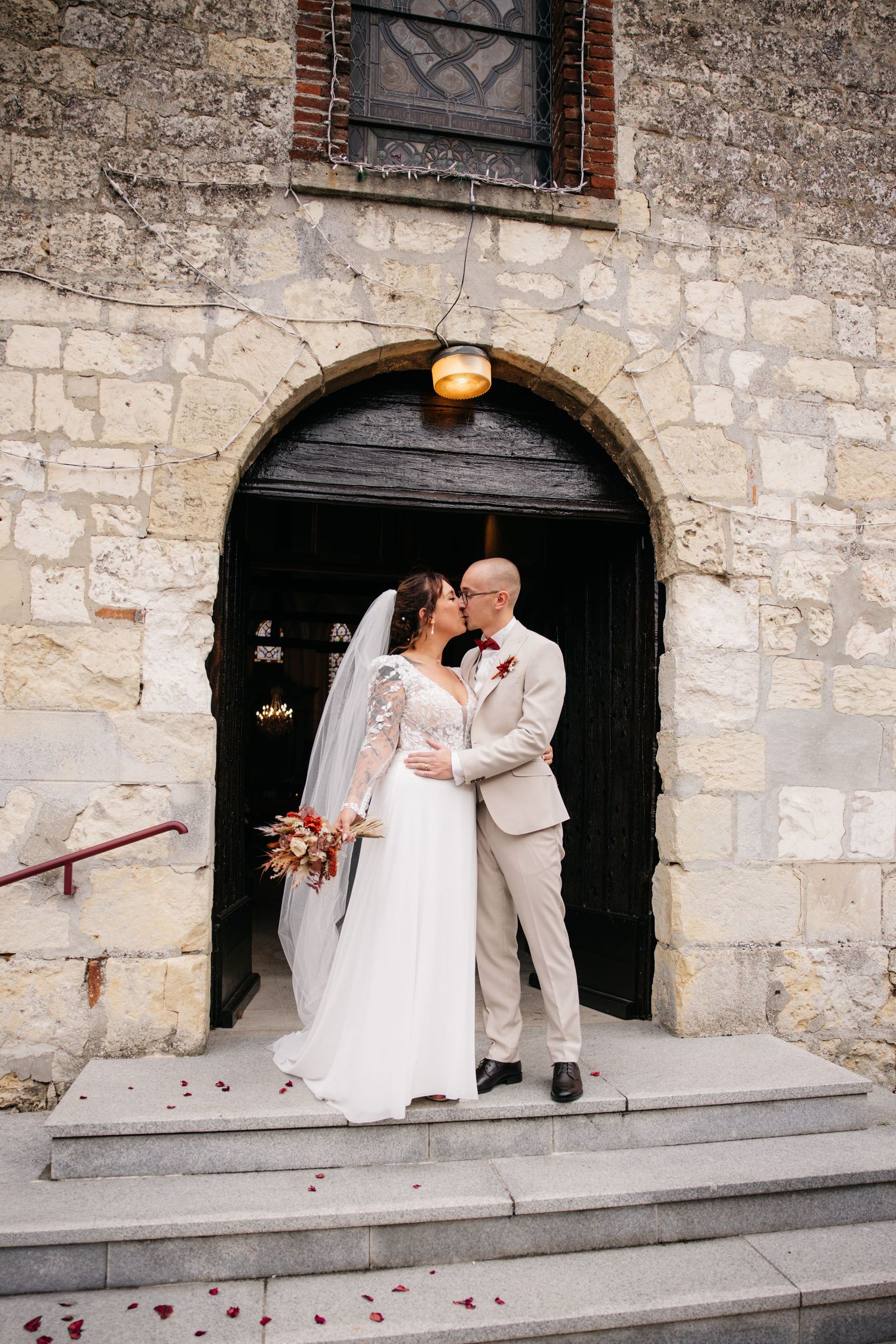 Les mariés partagent un moment romantique sur les marches en pierre d'une église, la mariée tenant un bouquet automnal dans sa robe blanche
