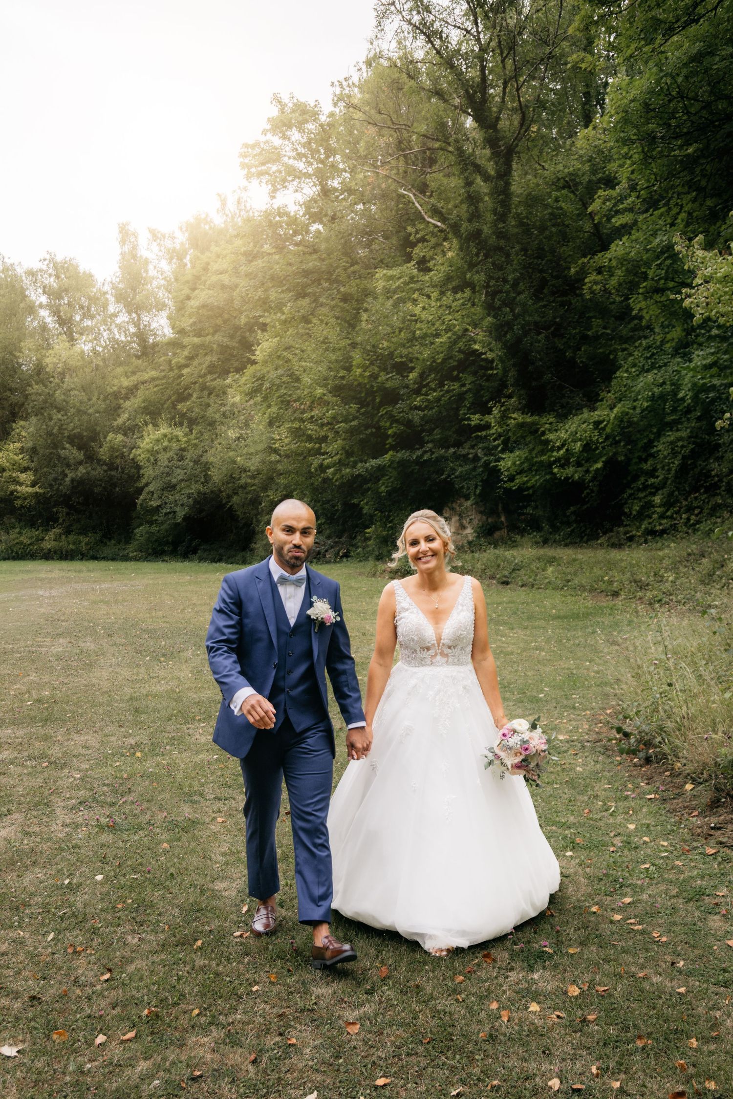 Une mariée souriante en robe blanche tient un bouquet tout en marchant avec son marié en costume bleu en extérieur.