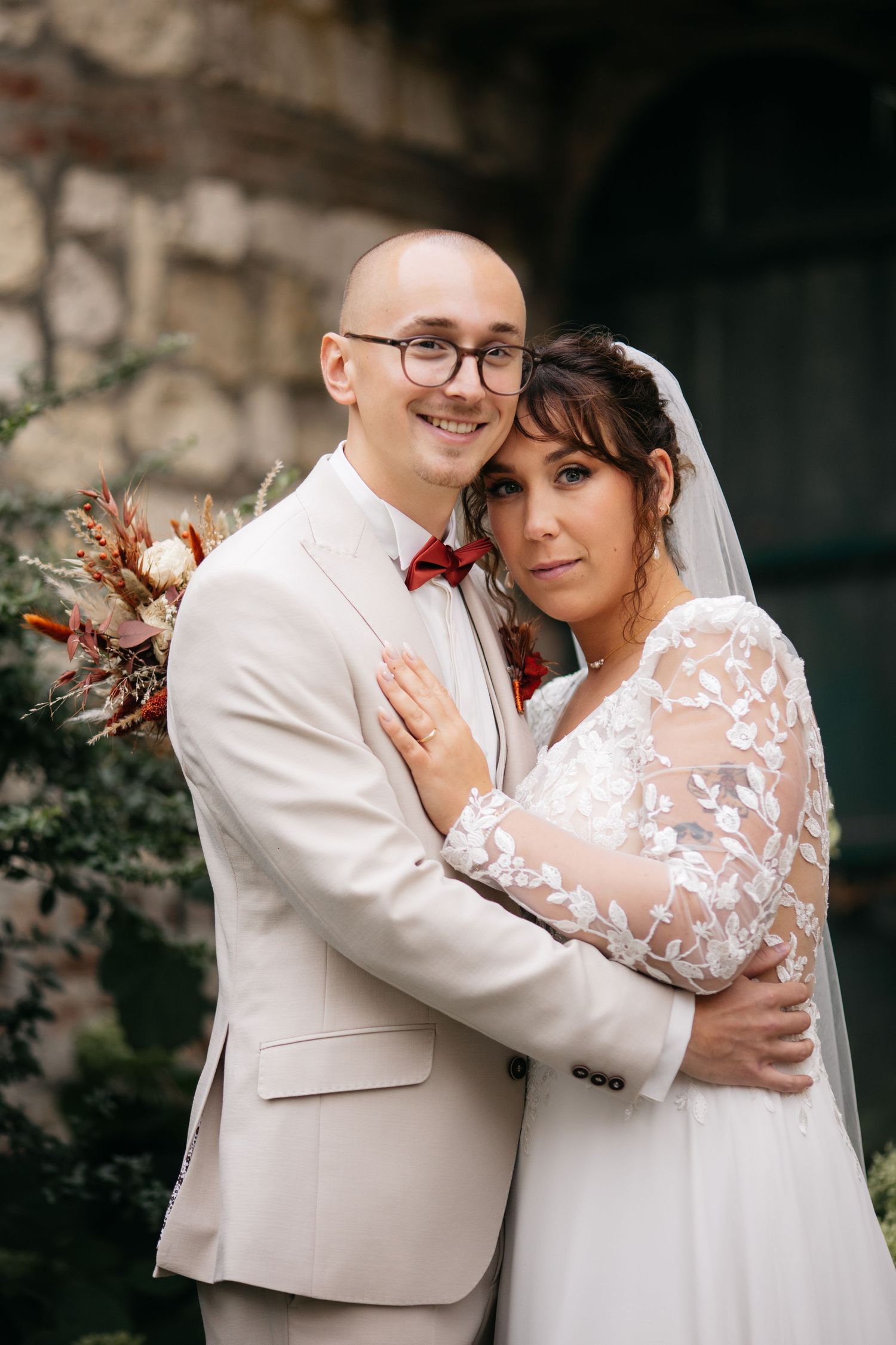 Un marié souriant, chauve, en costume beige, enlace sa mariée vêtue d’une robe de mariée en dentelle et d’un voile à l’extérieur.