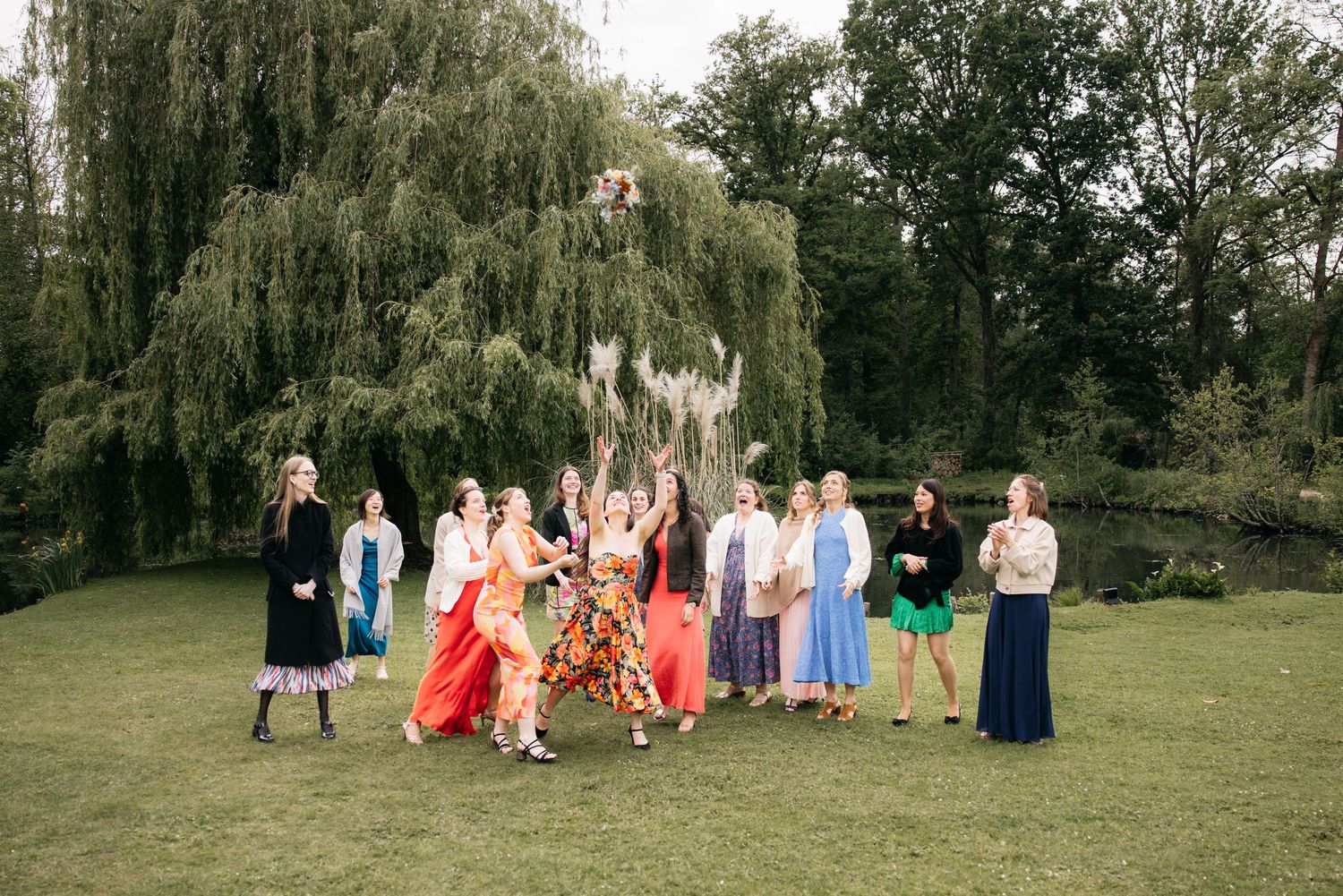 Groupe de femmes en robes colorées célébrant en extérieur, lançant un bouquet près d'un saule pleureur.