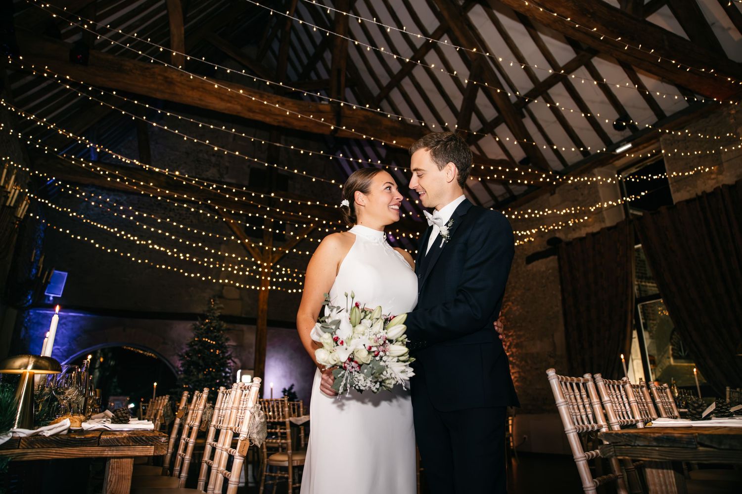 Mariés partageant un moment romantique dans une grange de mariage décorée de guirlandes lumineuses et de chaises en bois rustique.