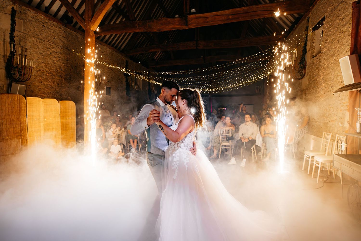 Les mariés partagent leur première danse dans une grange rustique, avec guirlandes lumineuses et effet de brume de glace carbonique.