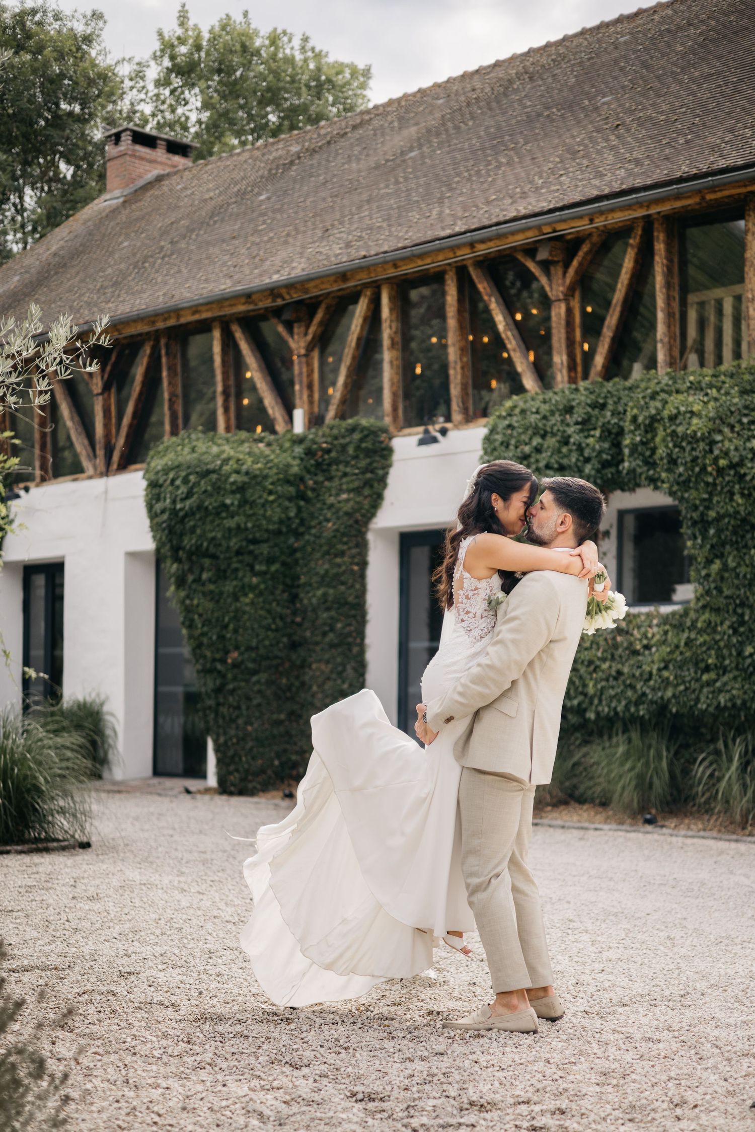 Le marié soulève la mariée en robe blanche devant un lieu champêtre recouvert de lierre, sur un chemin de gravier, et échange un baiser