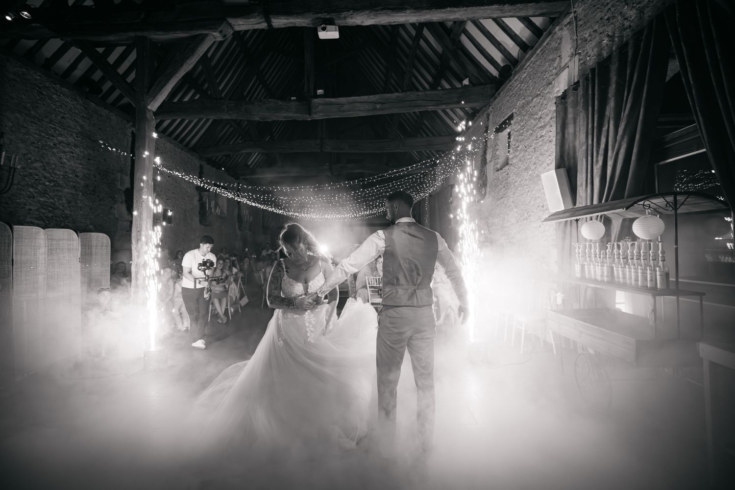 Photo en noir et blanc des mariés partageant leur première danse dans une grange rustique, avec guirlandes lumineuses et brume théâtrale.
