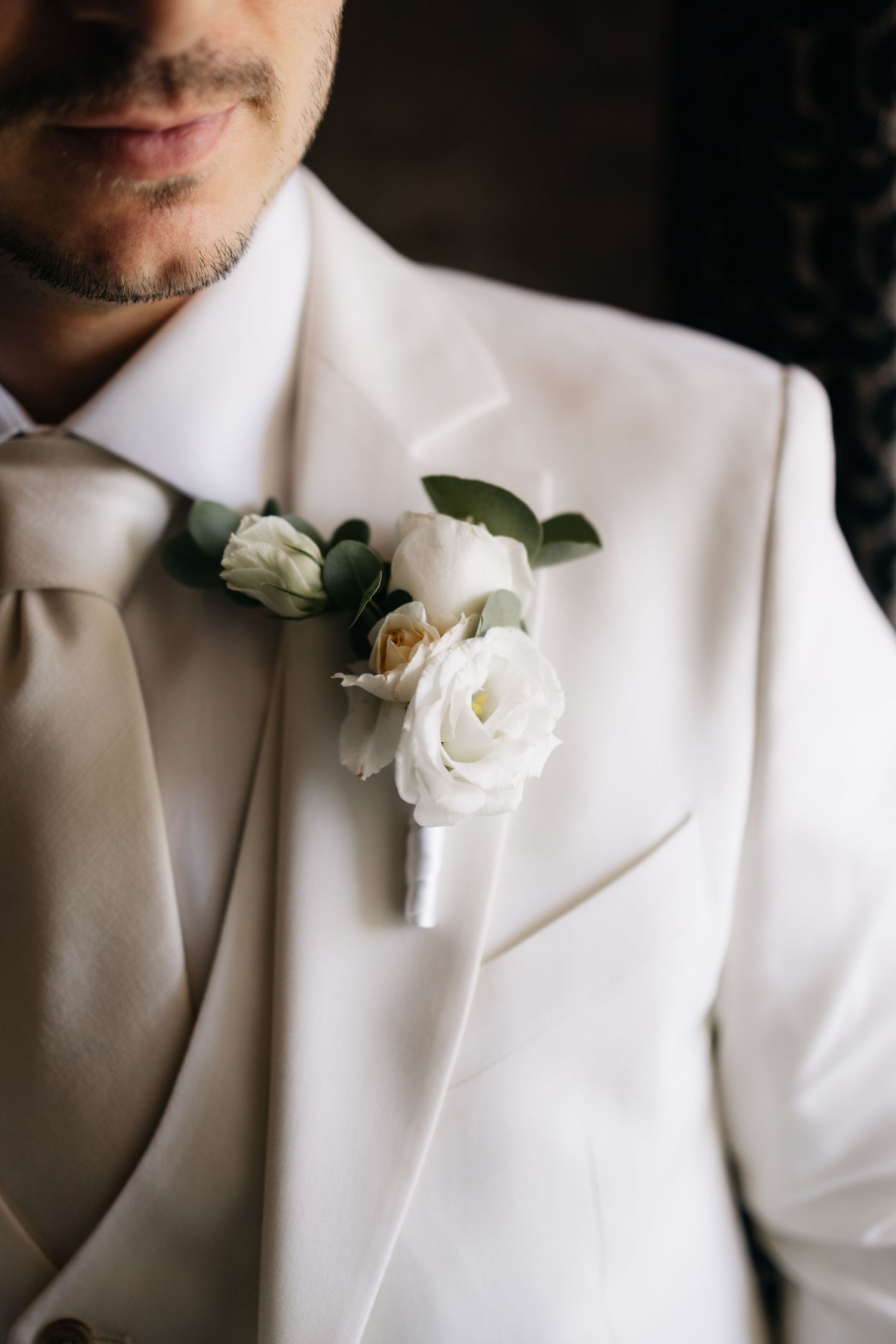 Gros plan sur un marié en costume blanc, avec une boutonnière de rose blanche et de la verdure de sauge sur le revers.