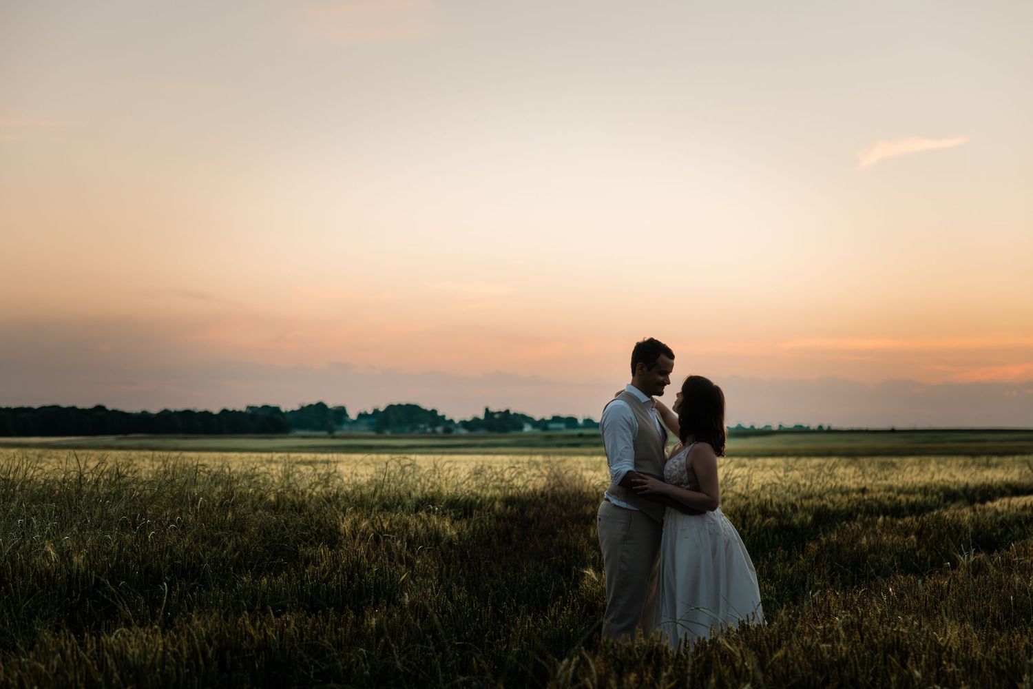 Un couple s'embrasse dans une robe blanche et des vêtements de cérémonie alors qu'il se tient dans un champ de blé au coucher du soleil