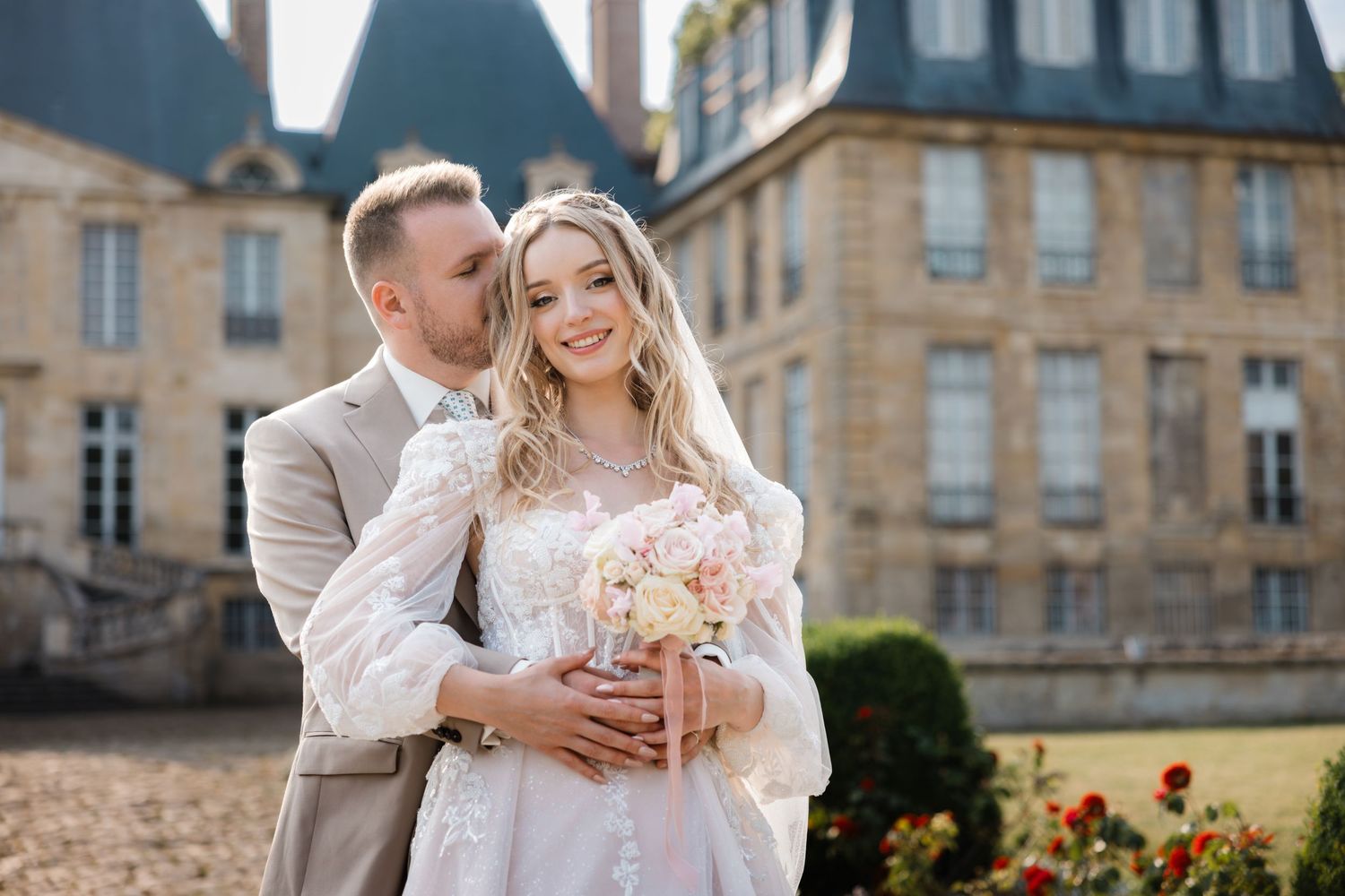 A couple embraces in formal wedding attire outside a majestic French chateau with pink floral bouquet.