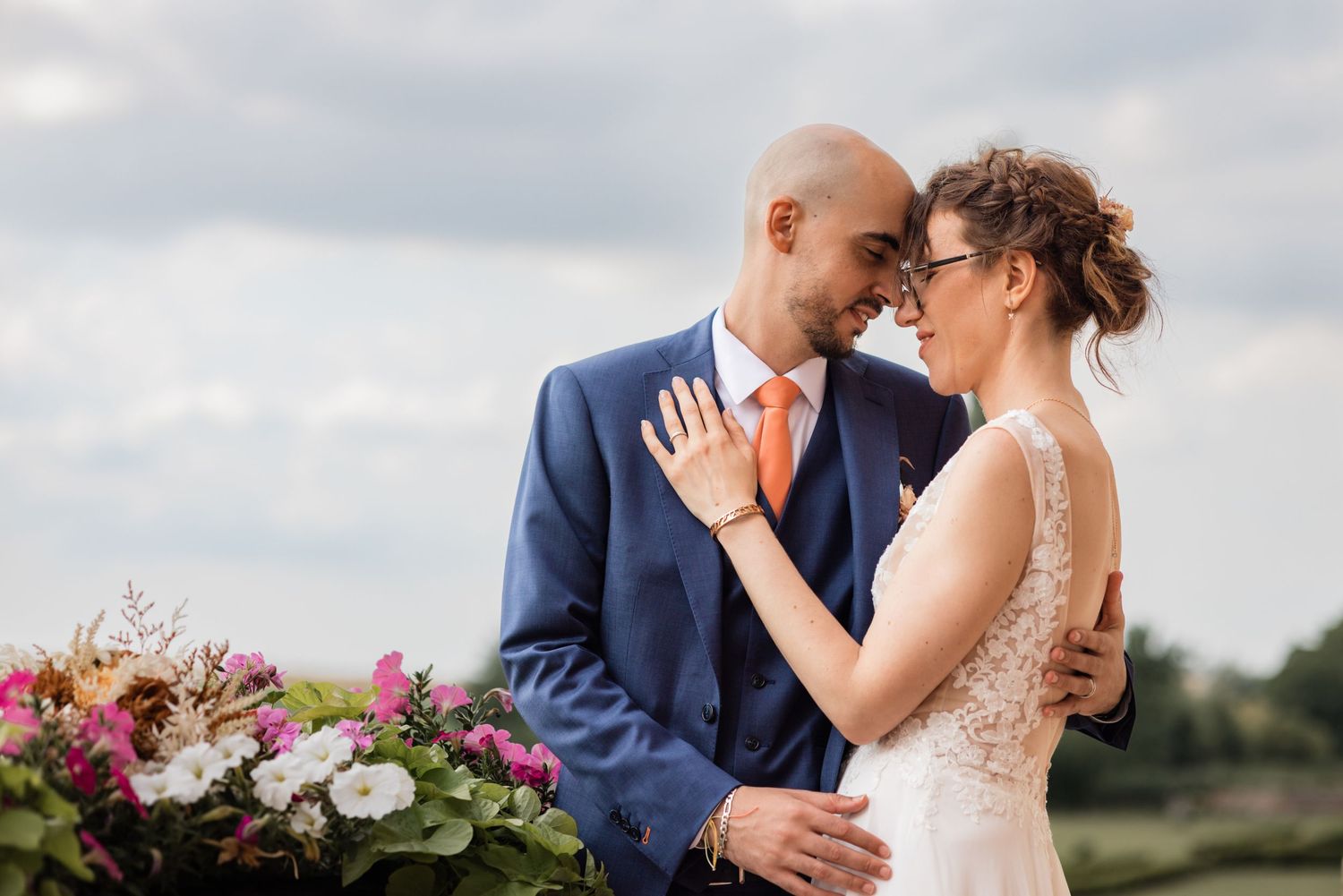 Un tendre moment de mariage capturé à l'extérieur avec des arrangements floraux de fleurs roses et blanches contre un ciel nuageux.