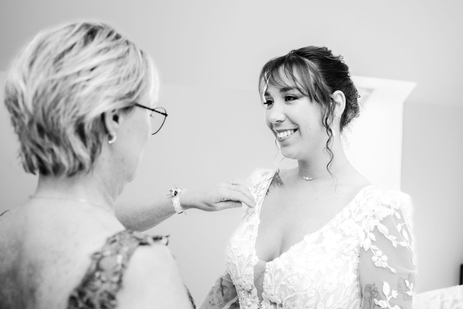 Photo en noir et blanc d'une mariée en robe de dentelle souriante tandis qu'une femme plus âgée lui touche l'épaule pendant les préparatifs