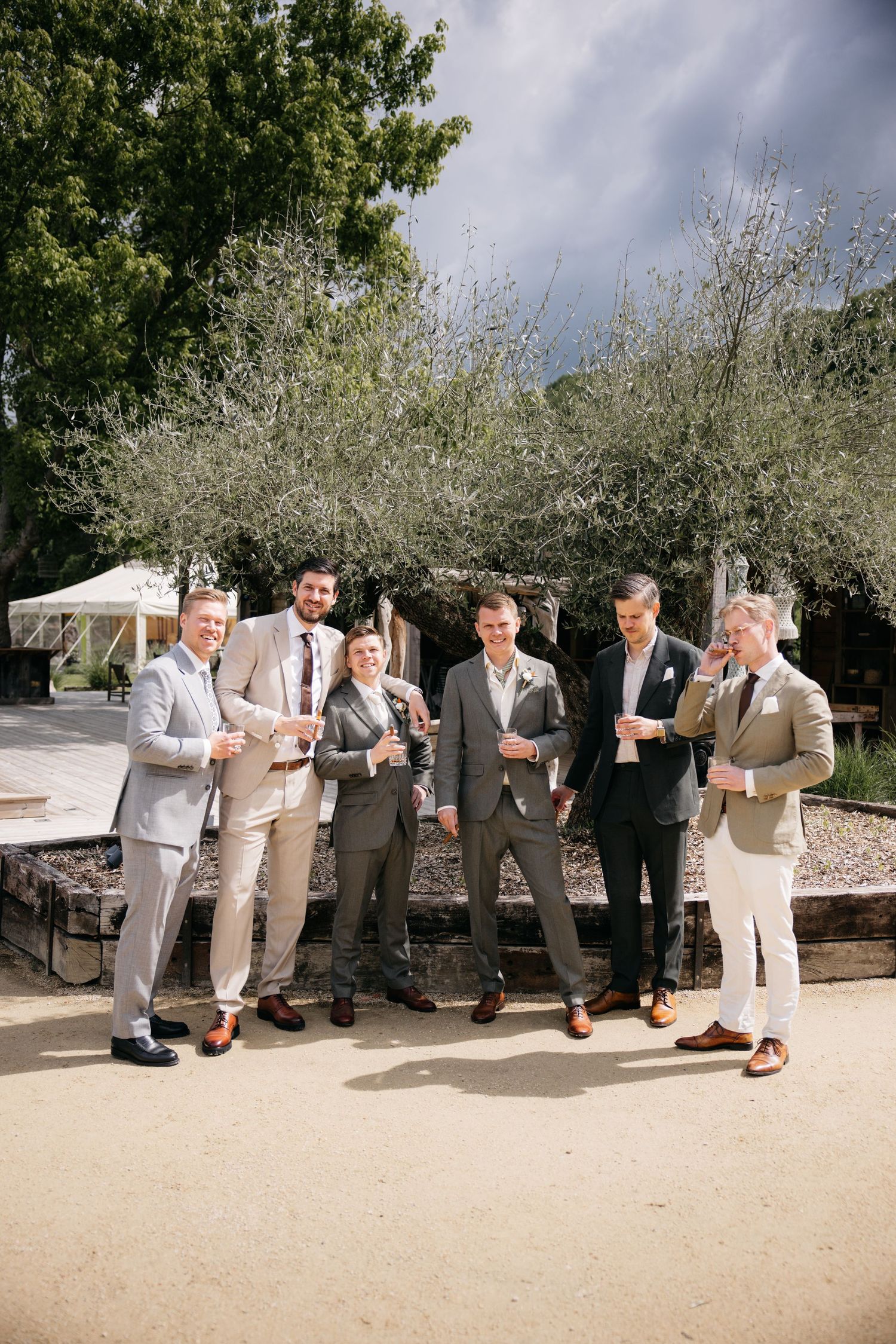 Six garçons d'honneur en costume tenant des verres posent en extérieur dans un lieu de mariage entouré d'arbres.