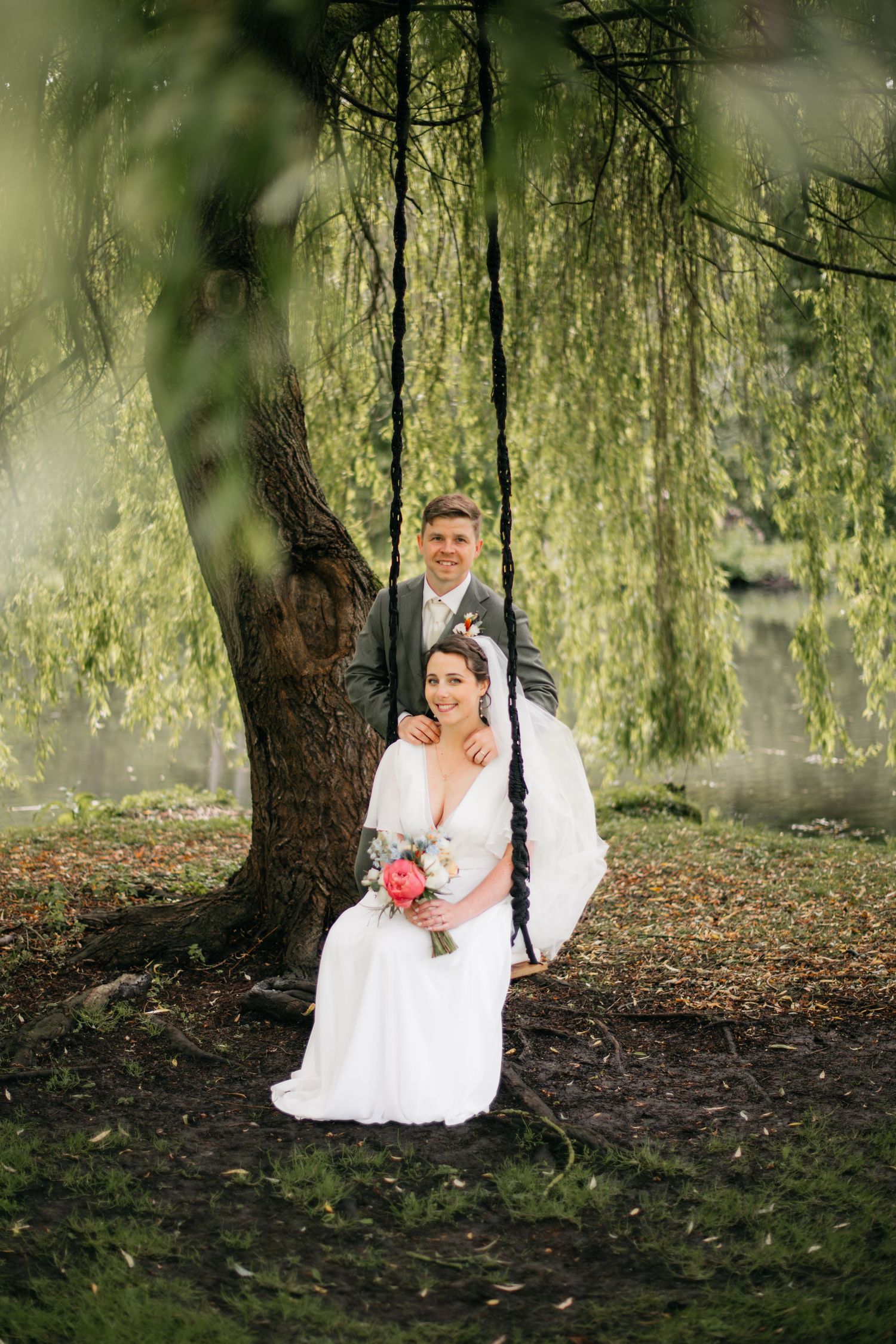 La mariée assise sur une balançoire tenant un bouquet, le marié debout derrière elle, sous un saule pleureur.
