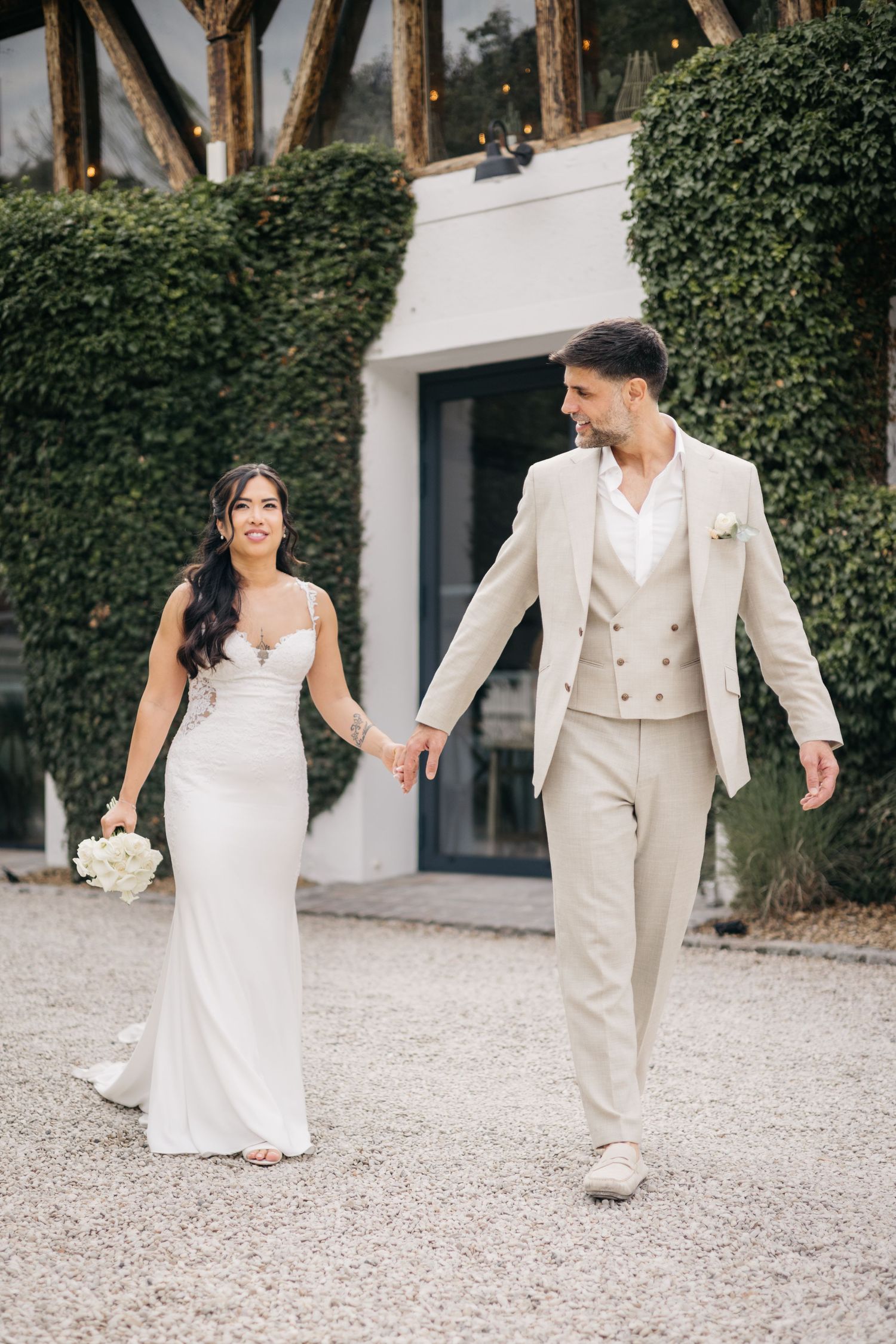 Un couple de mariés se tient la main et marche à l'extérieur, elle en robe blanche, lui en costume beige, avec de la verdure en arrière-plan