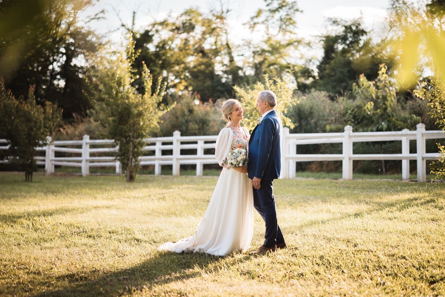 A couple in wedding attire embraces in a sunlit field with white wooden fences on a golden afternoon.