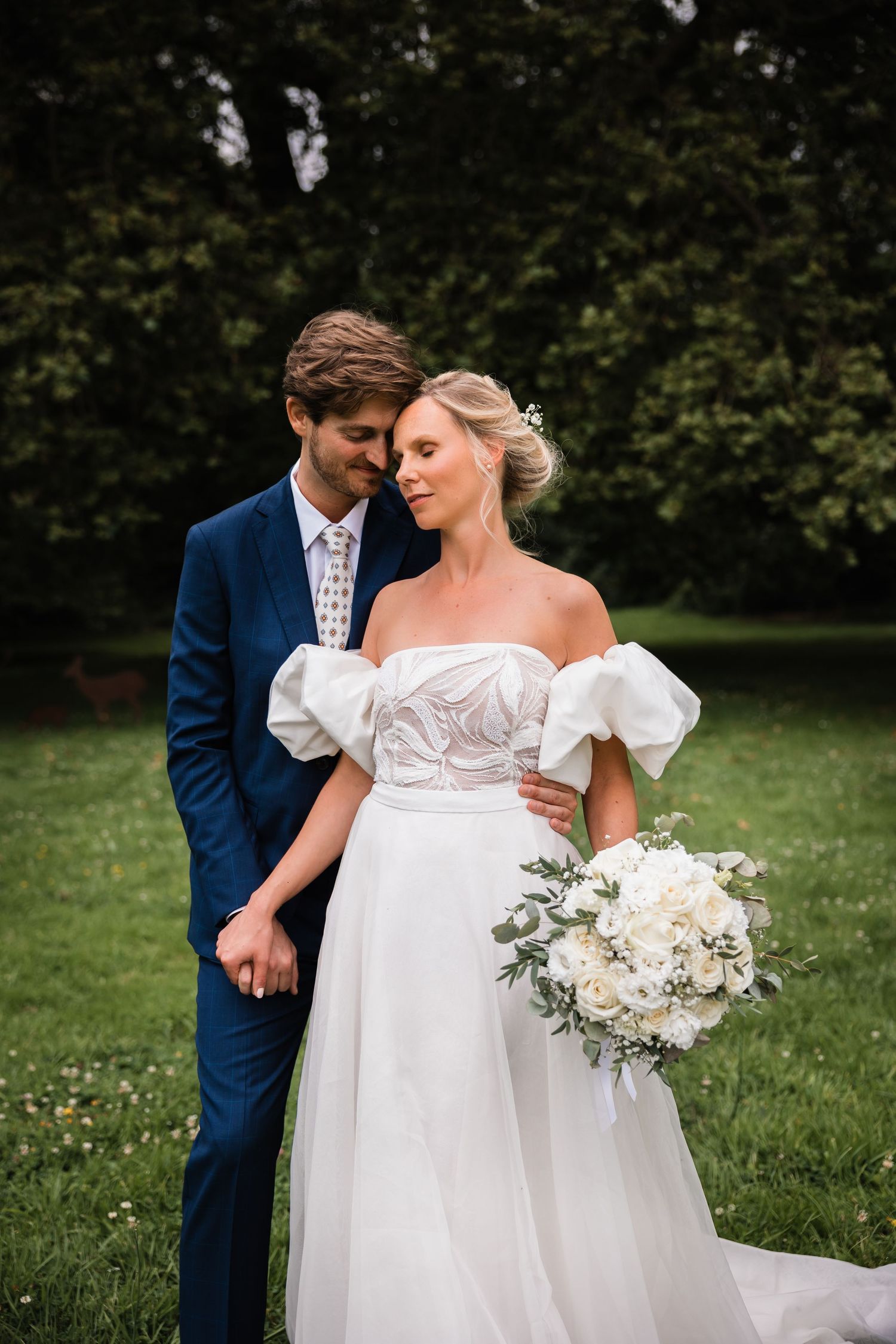 Un couple pose à l'extérieur le jour de leur mariage avec la mariée dans une robe à manches bouffantes tenant un bouquet de fleurs blanches.