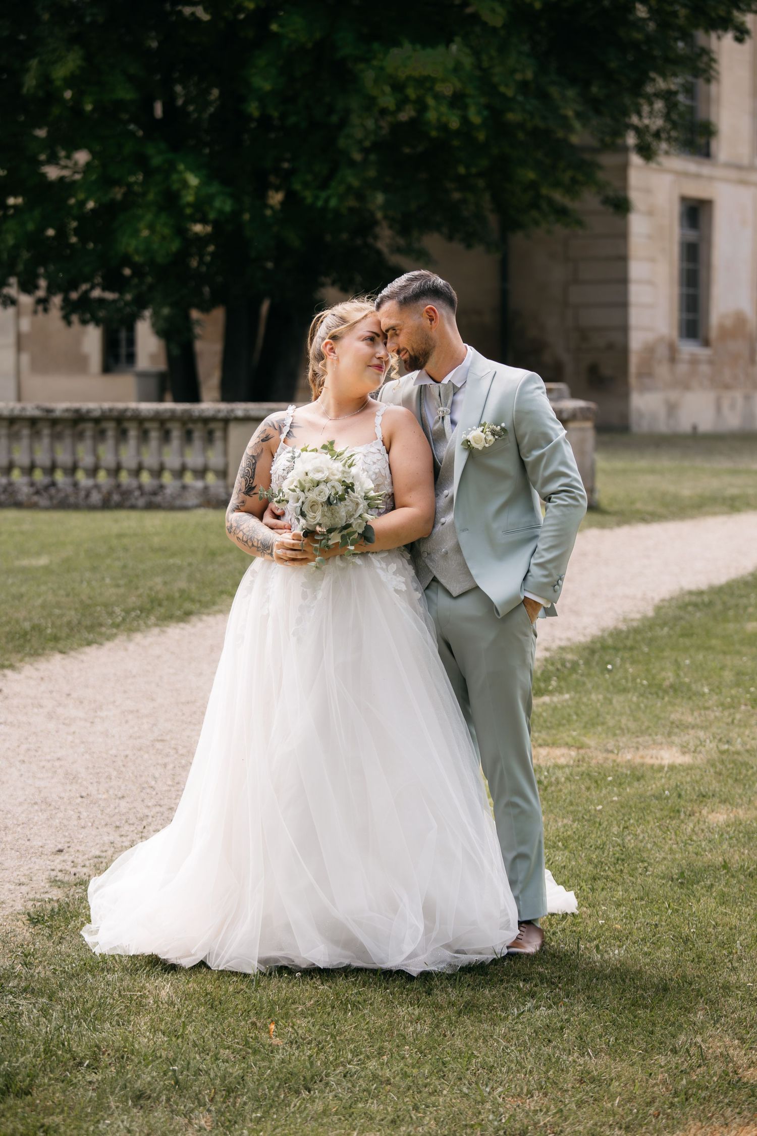 Mariée en robe de tulle blanche tenant un bouquet aux côtés du marié en costume sauge, posant en extérieur près d'un bâtiment historique.