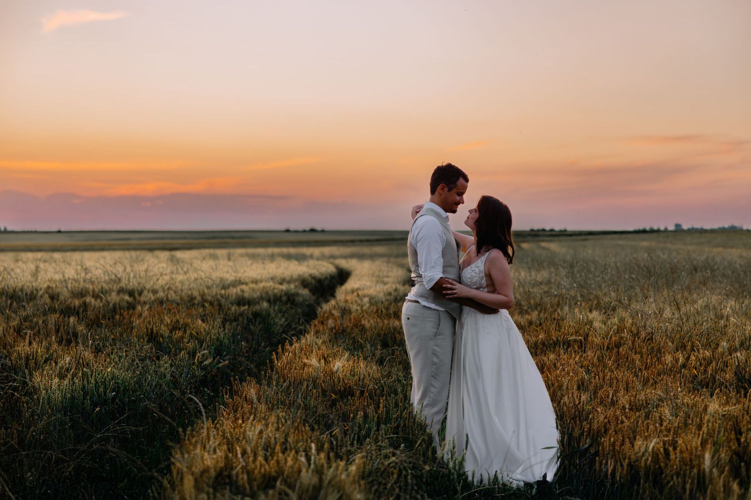 Un couple en tenue de mariage s'embrasse dans un champ de blé doré au coucher du soleil avec un ciel orange derrière eux.