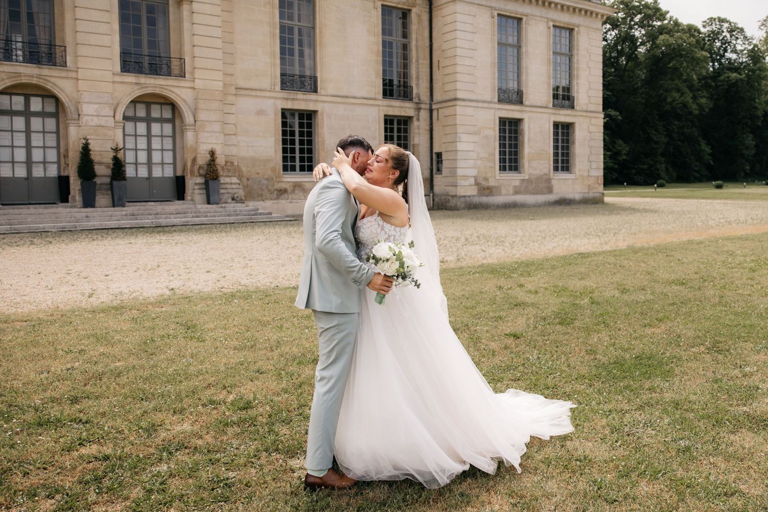 Les mariés échangent un baiser romantique devant un élégant château français, la mariée tenant un bouquet blanc.